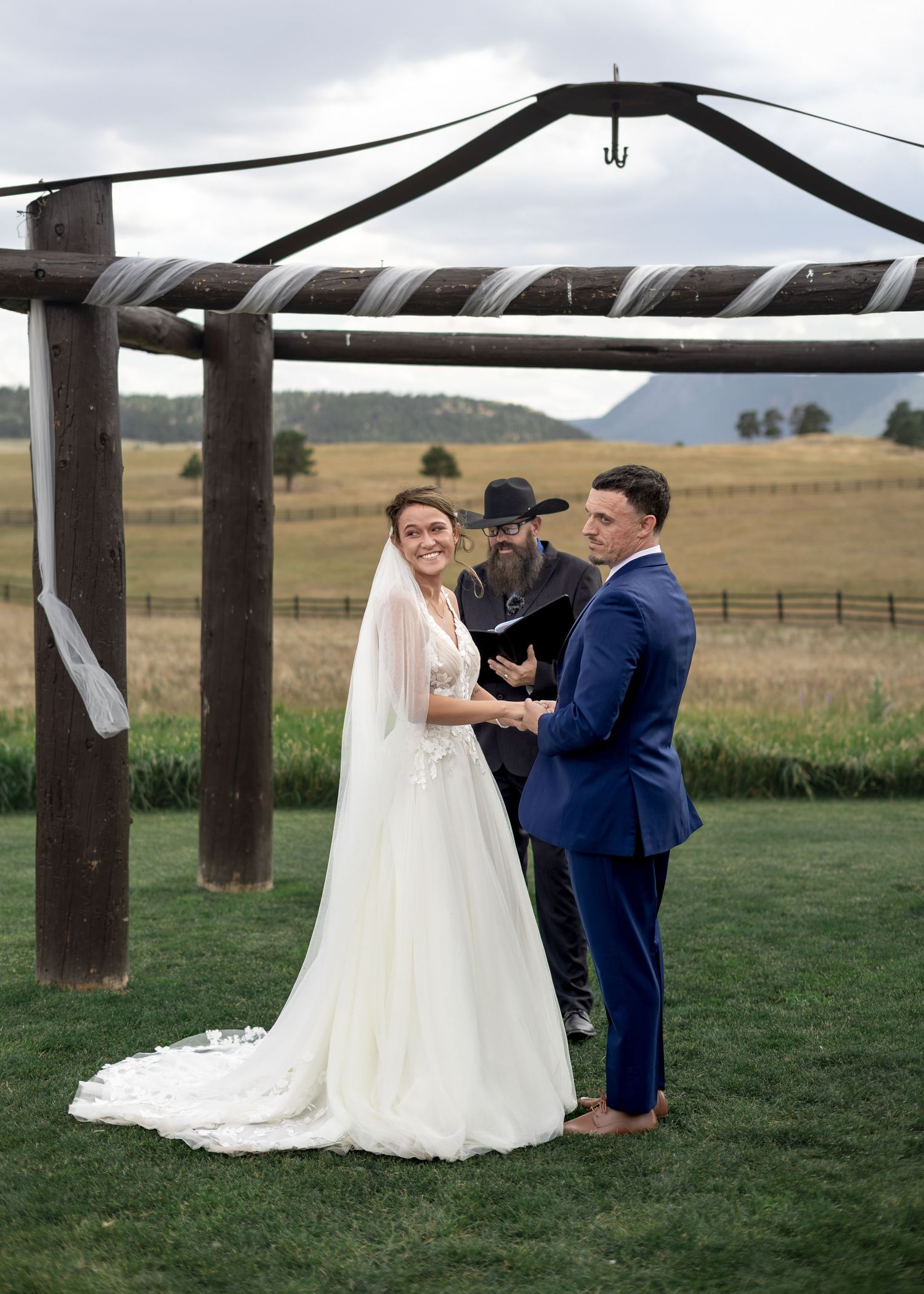 Bride and groom exchange vows under rustic wooden arbor at outdoor ranch wedding ceremony with mountain views.