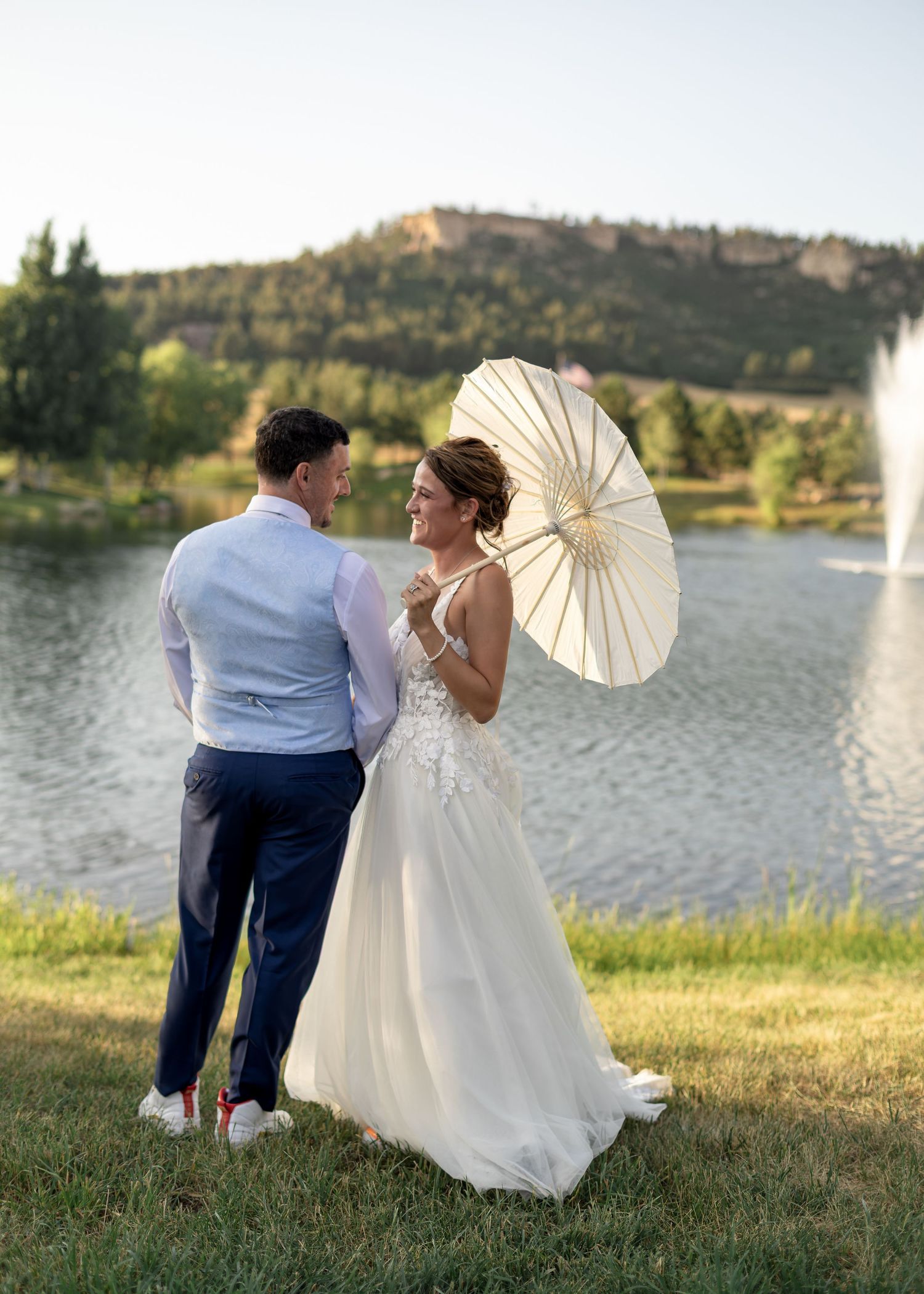 Bride holding a white parasol smiling at groom near a lake with mountain backdrop.