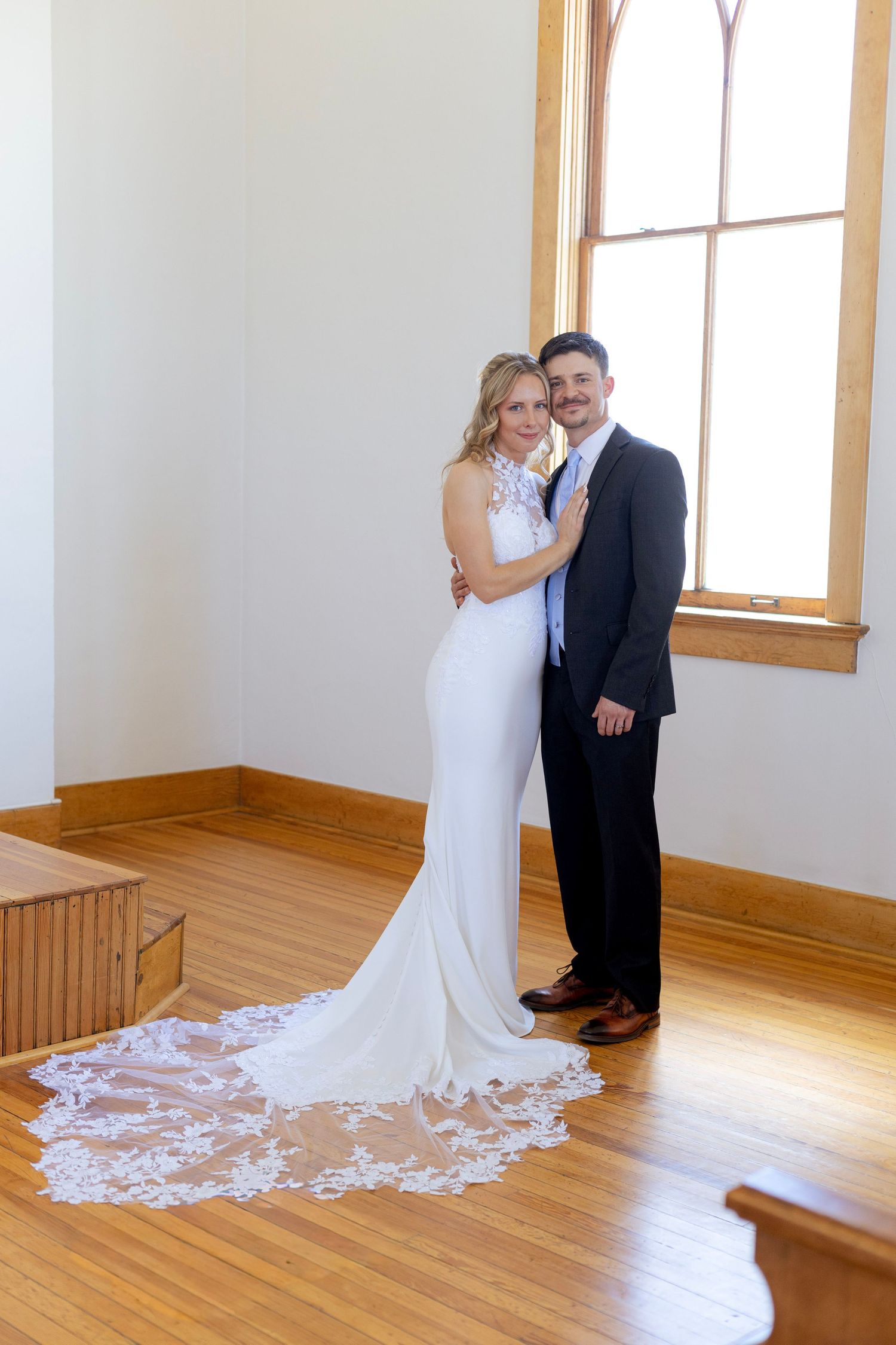Bride and groom pose together by window in chapel, brides lace train spread on floor.