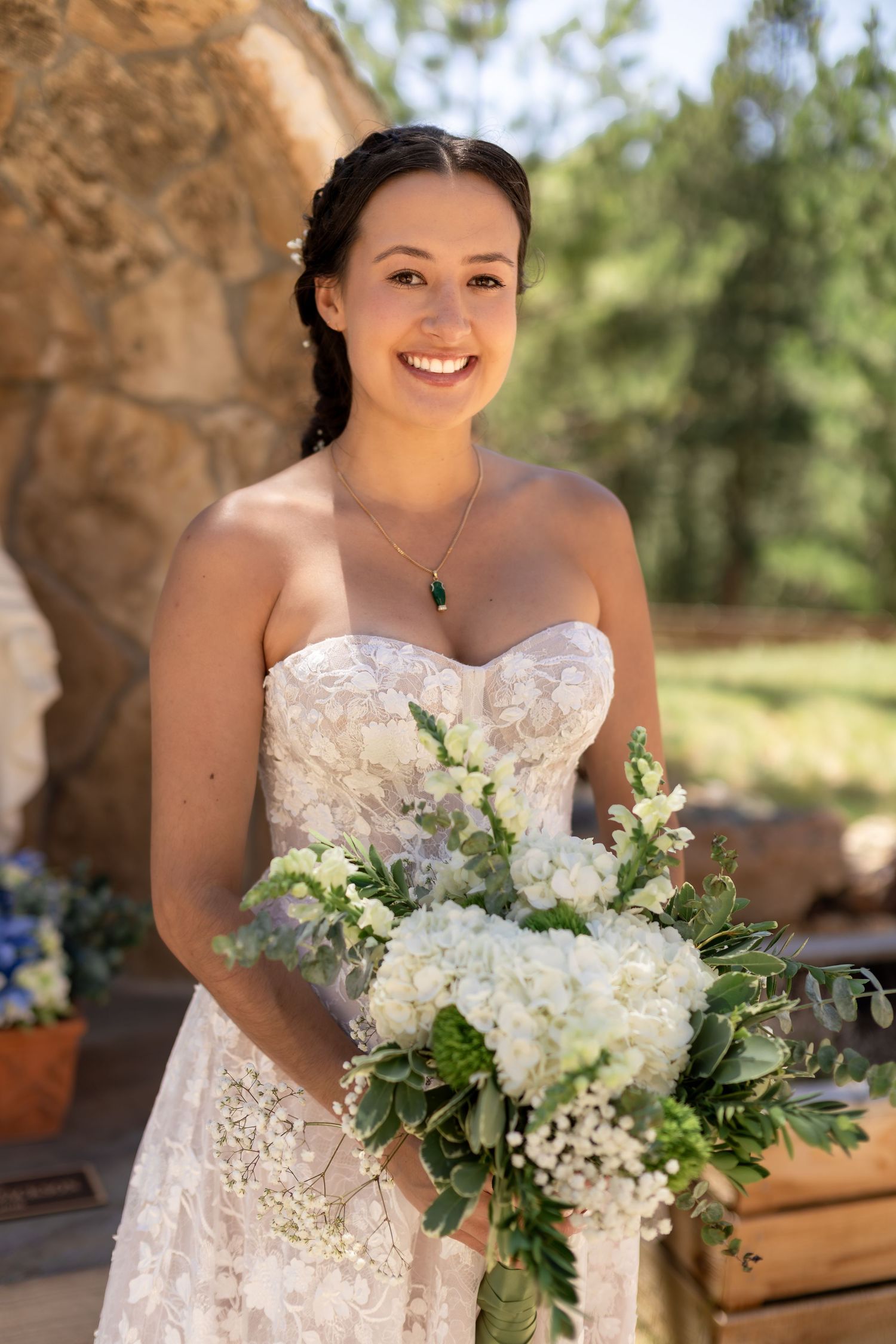 Smiling bride in strapless lace gown holding white hydrangea bouquet with greenery outdoors.