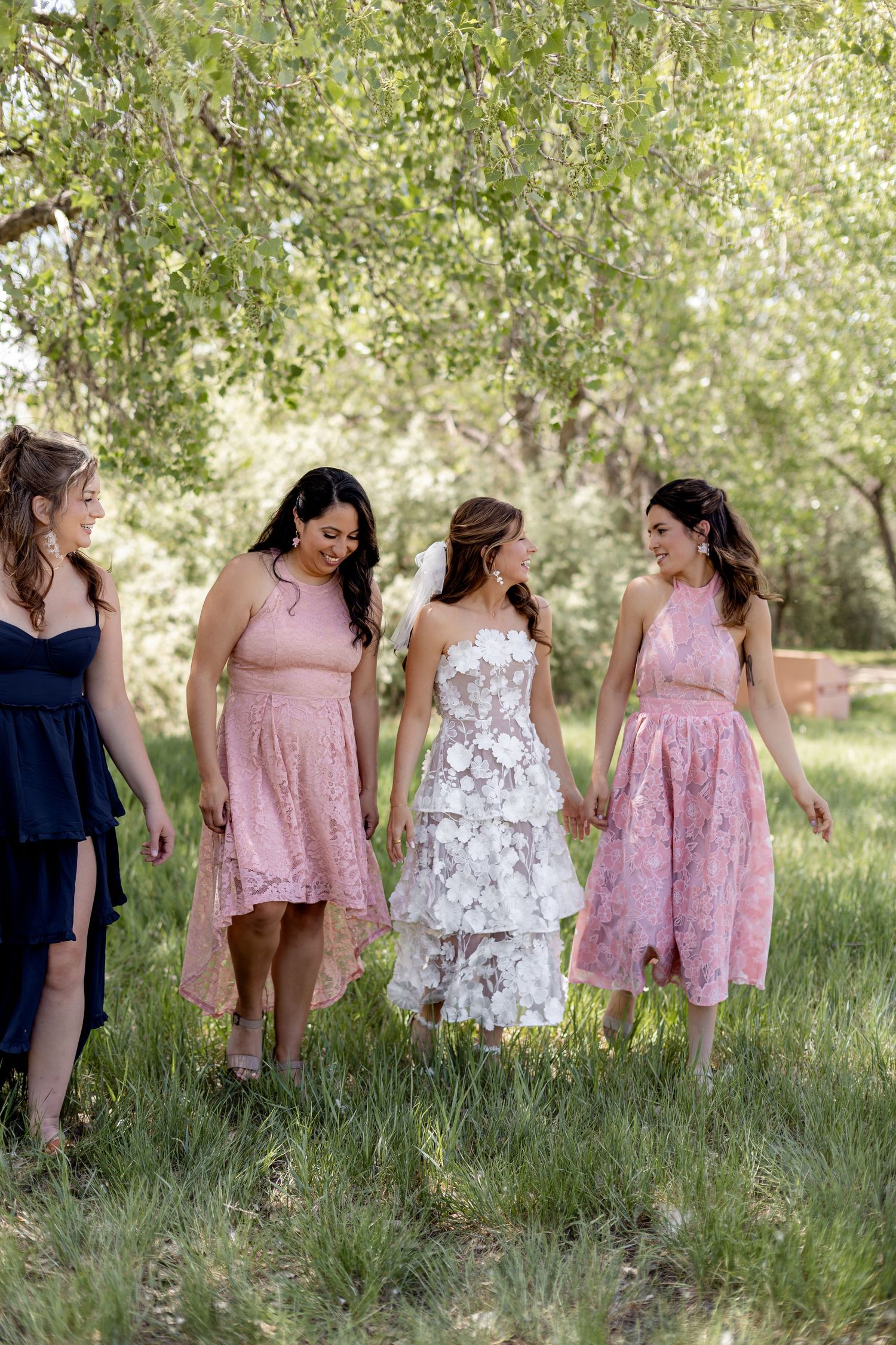 Bride in floral white gown walks with three bridesmaids in pink and navy dresses outdoors.
