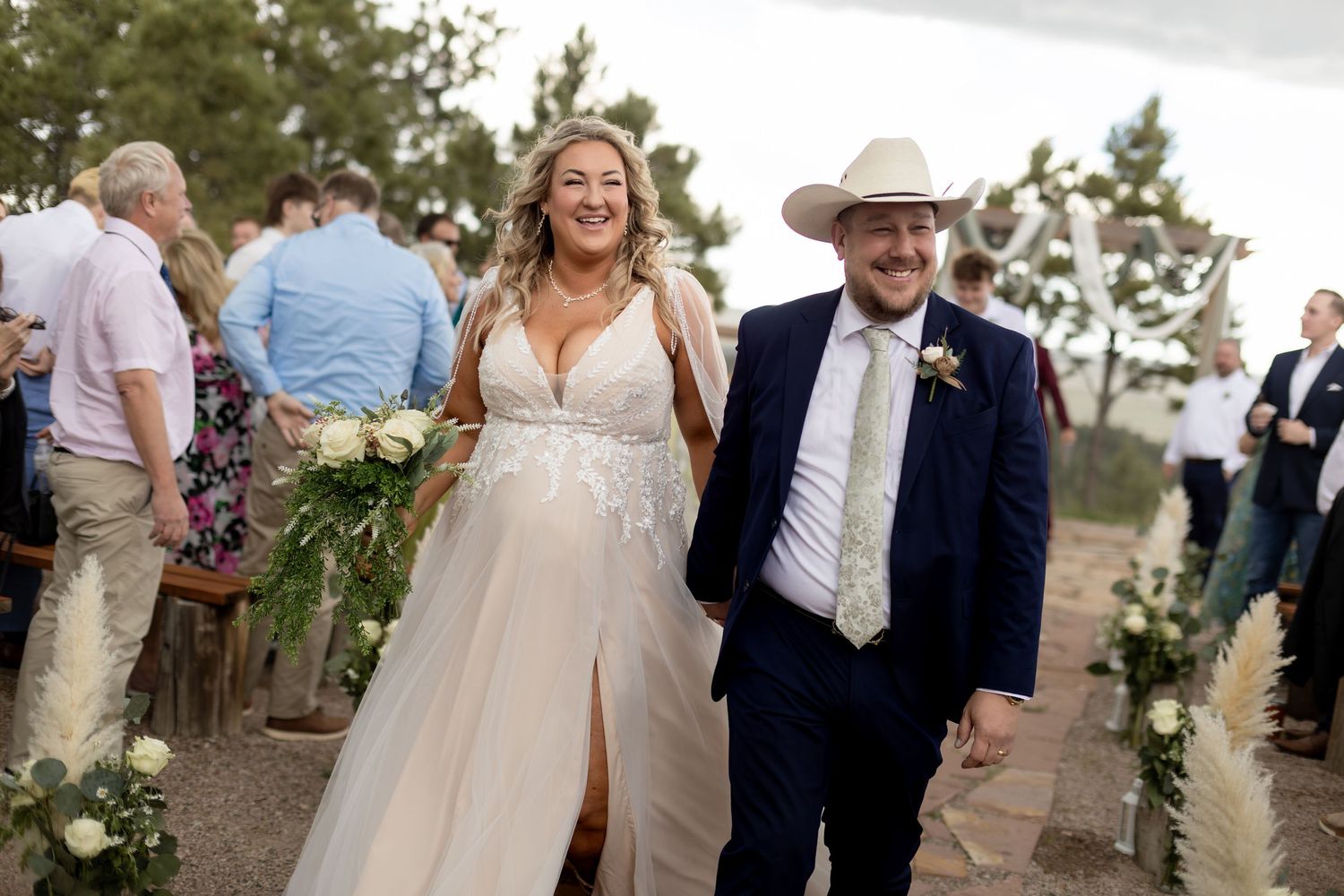 Newlywed couple walking down the aisle, bride in lace gown and groom in navy suit with cowboy hat.