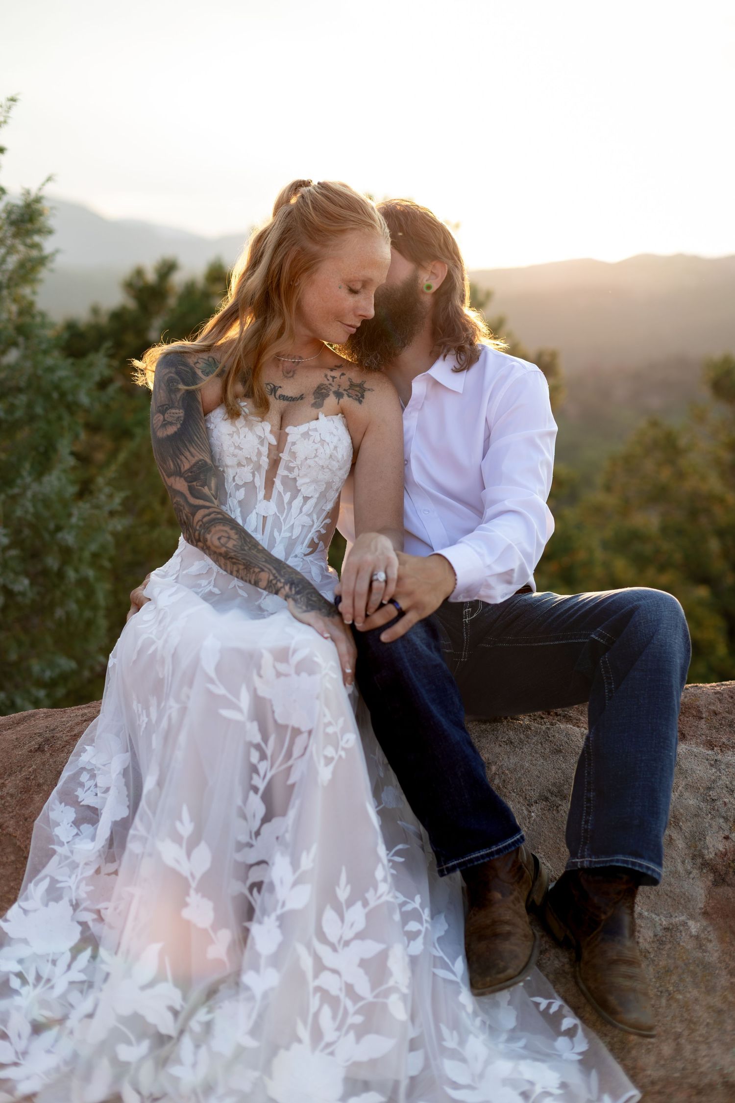 Tattooed bride in lace gown sits with bearded groom on rocks at golden hour sunset in Colorado.