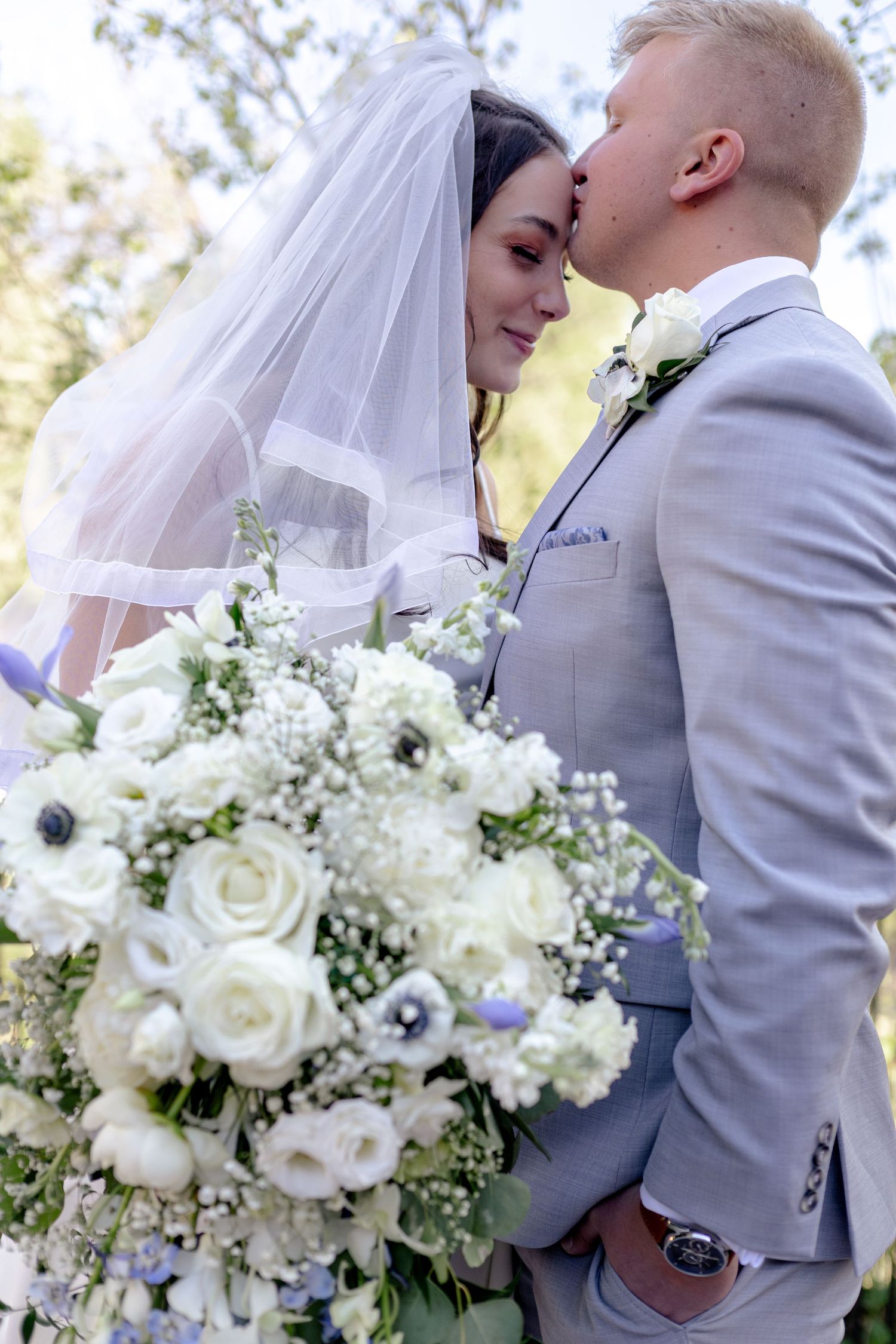 Groom in gray suit kisses bride wearing veil while holding lush white wedding bouquet outdoors.