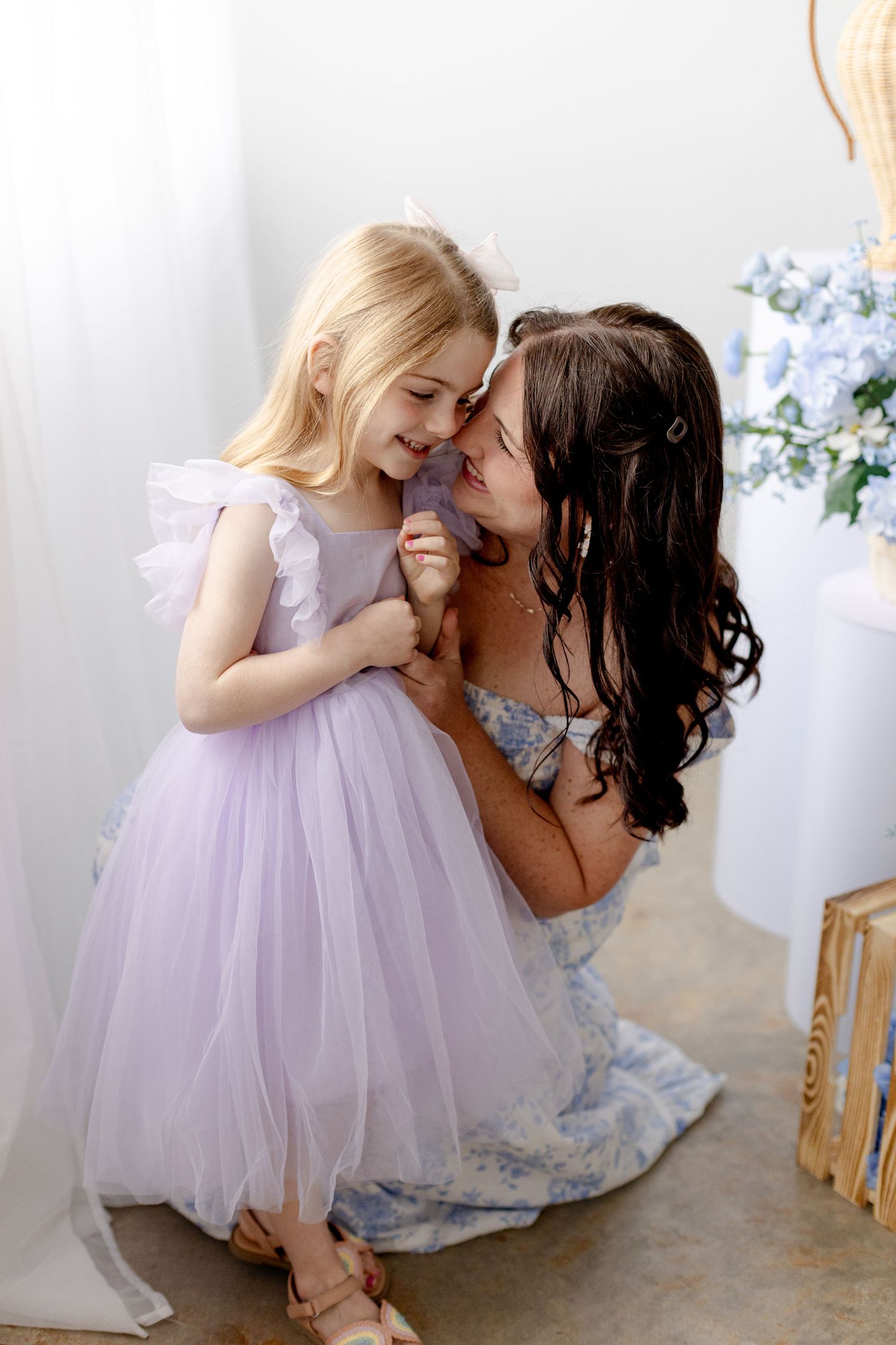 A mother in a floral dress shares a tender moment with her young blonde daughter in a lavender tulle gown.
