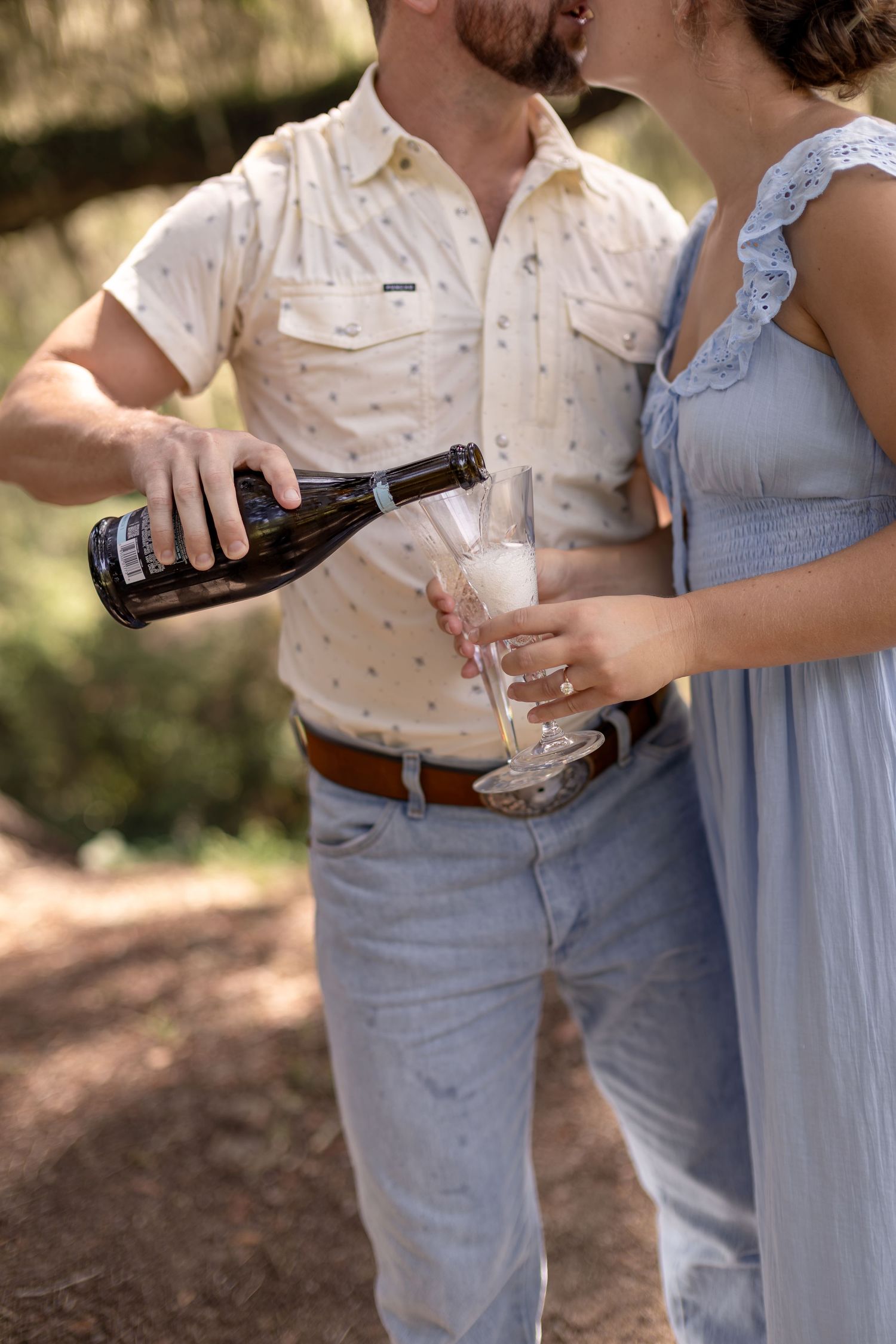 Man pours champagne into a flute glass while couple shares a kiss outdoors in nature.