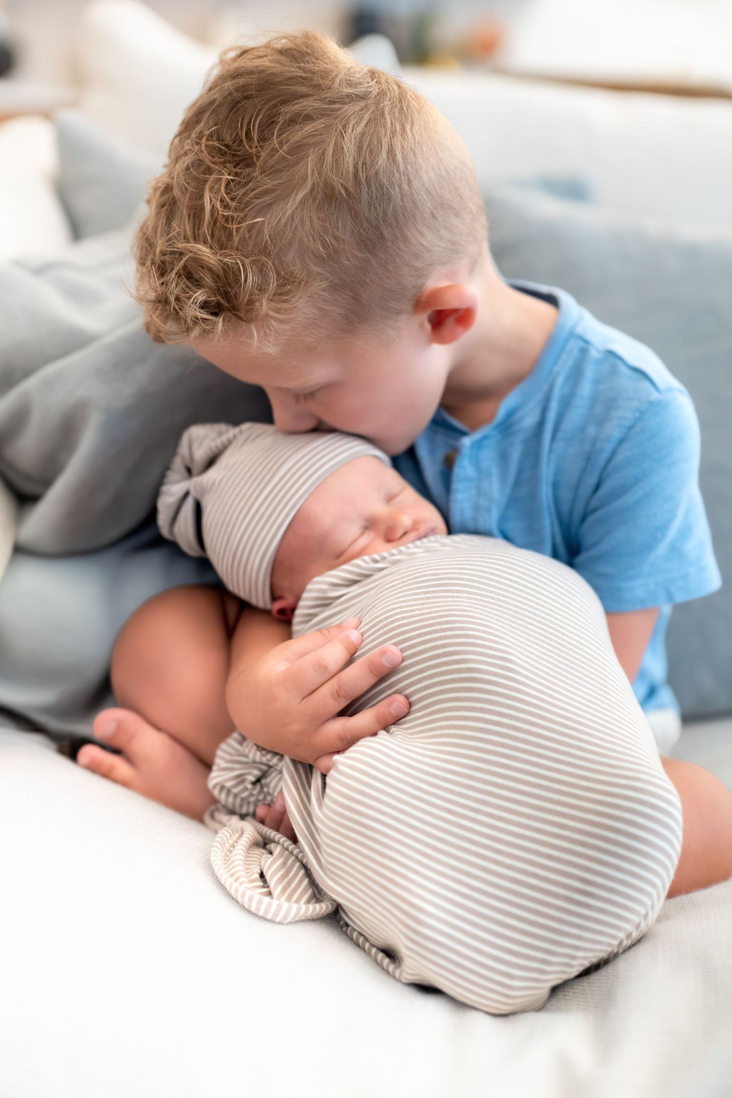 Young boy in blue shirt gently kisses sleeping newborn sibling wrapped in striped swaddle blanket.