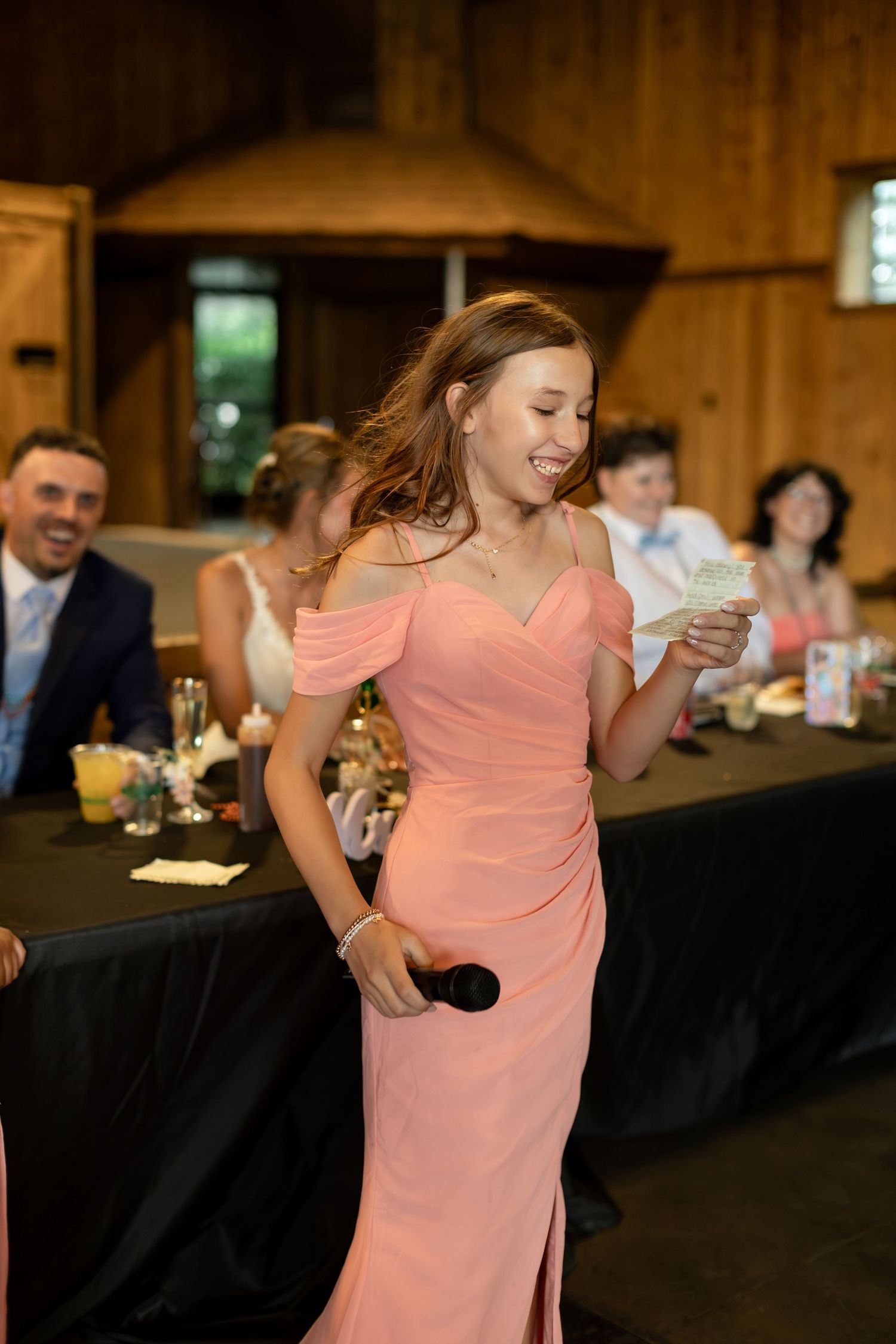 Bridesmaid in a coral off-shoulder gown delivers a heartfelt speech holding a microphone at a barn wedding reception.