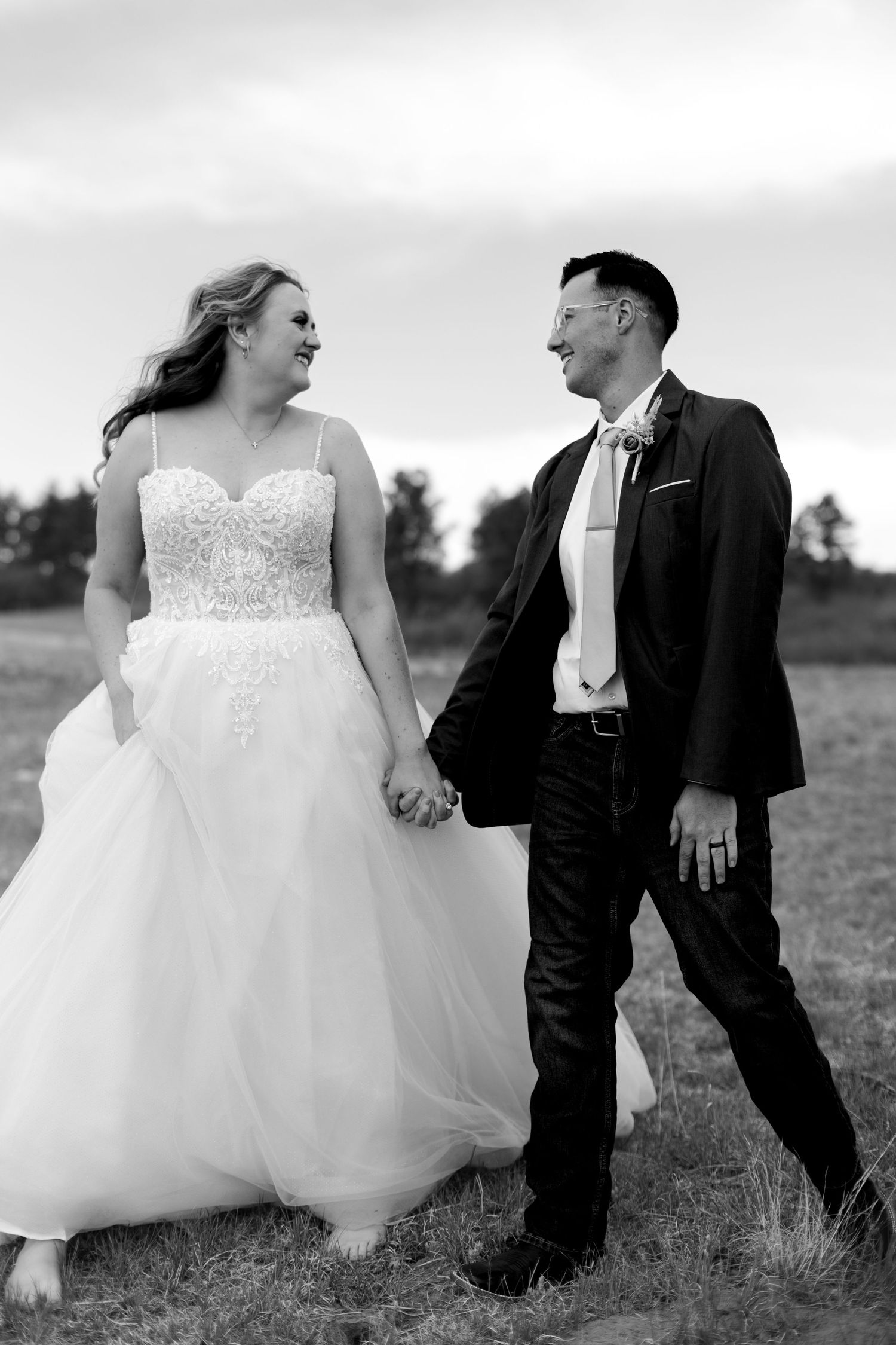Black and white photo of laughing bride and groom holding hands walking through open field.