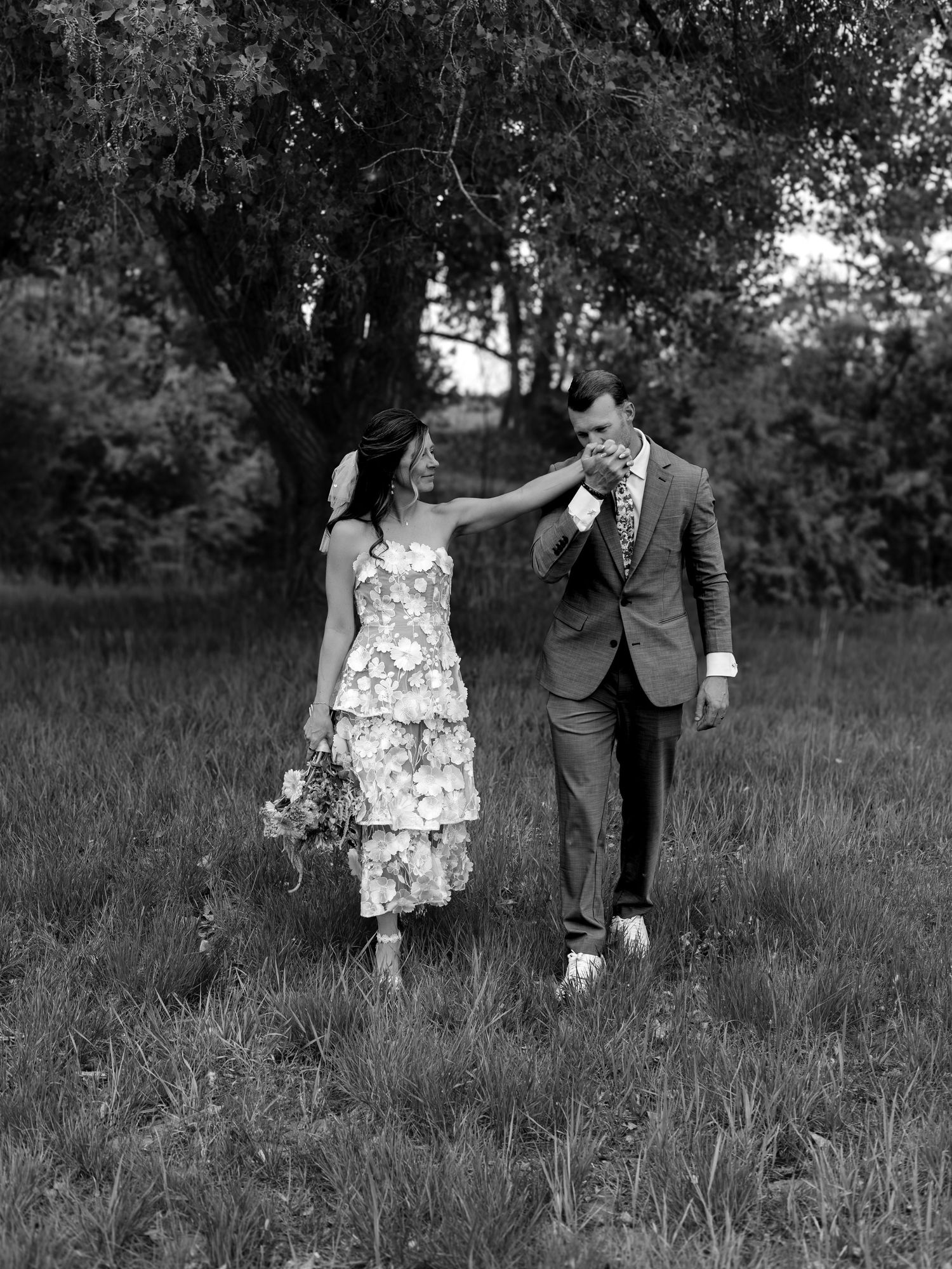 Black and white photo of a couple walking in a field, bride in floral dress holding bouquet, groom in suit kissing her hand.