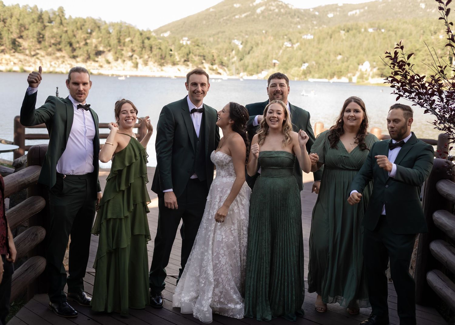 Wedding party poses and celebrates on lakeside deck with mountain scenery in background.