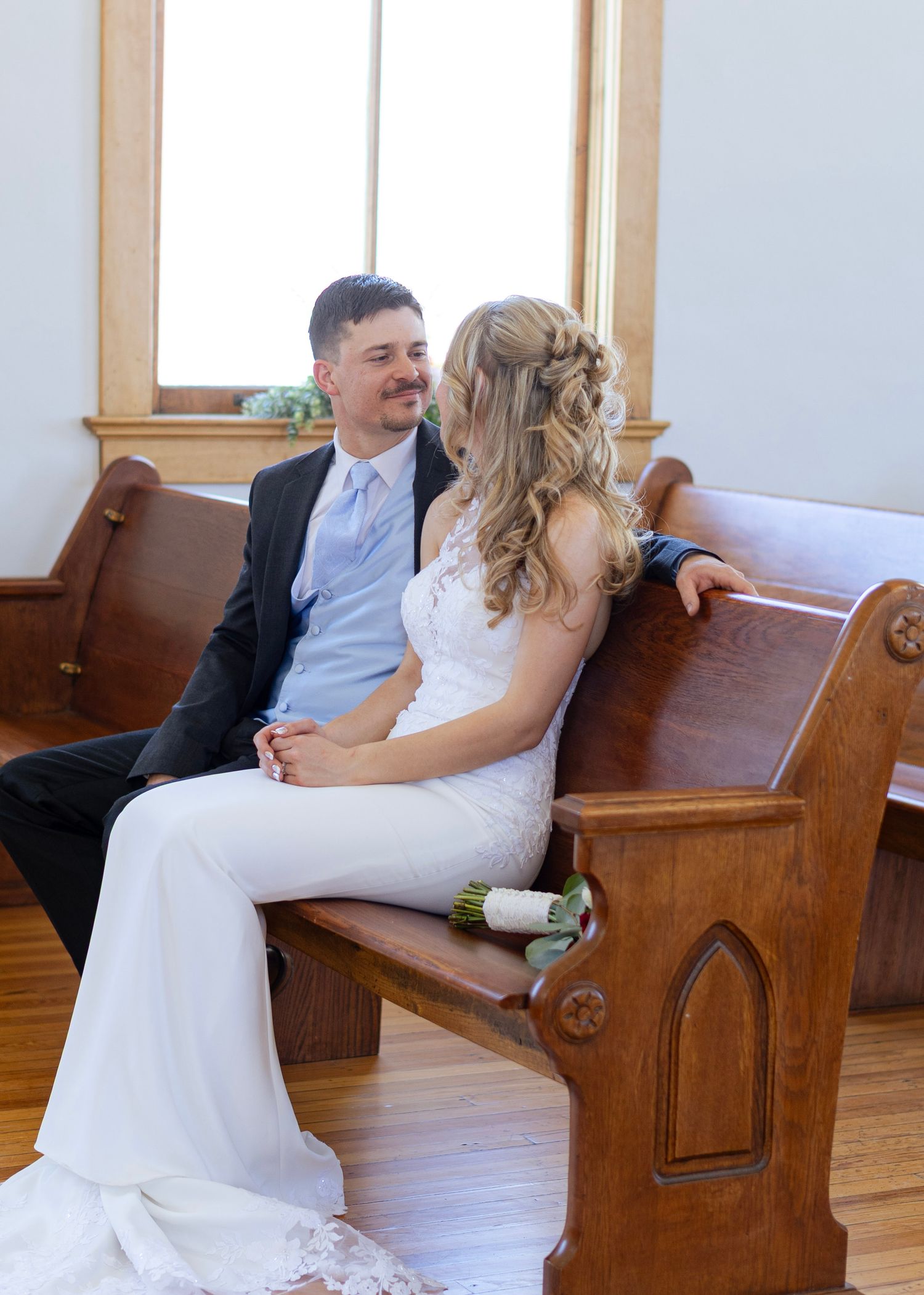 Bride and groom sitting together on wooden church pew gazing at each other on their wedding day.