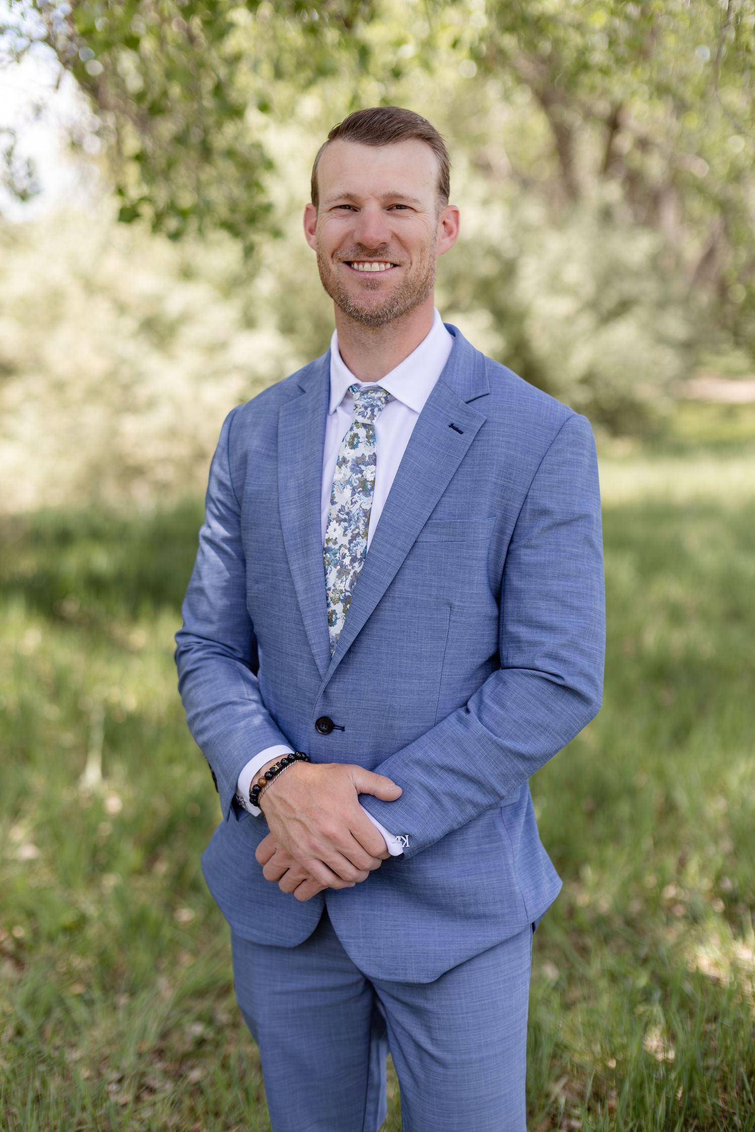 Handsome groom smiles in a light blue suit with floral tie at an outdoor wedding venue.