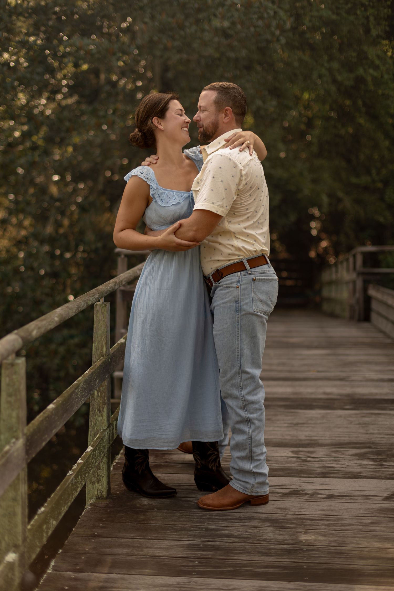 Couple embraces and smiles on rustic wooden bridge surrounded by lush green trees at dusk.