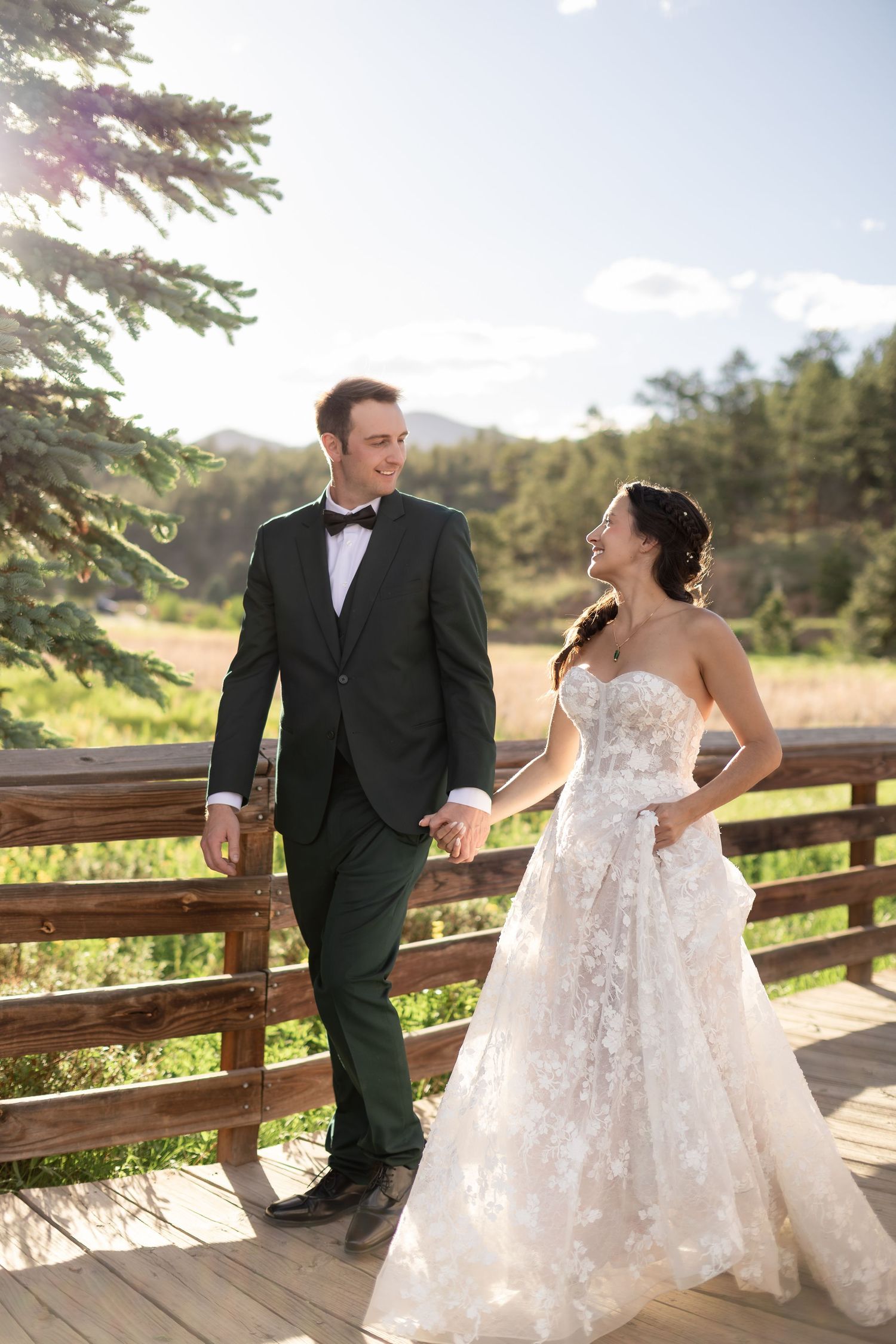 A bride and groom hold hands on a wooden deck surrounded by mountains and trees on their wedding day.