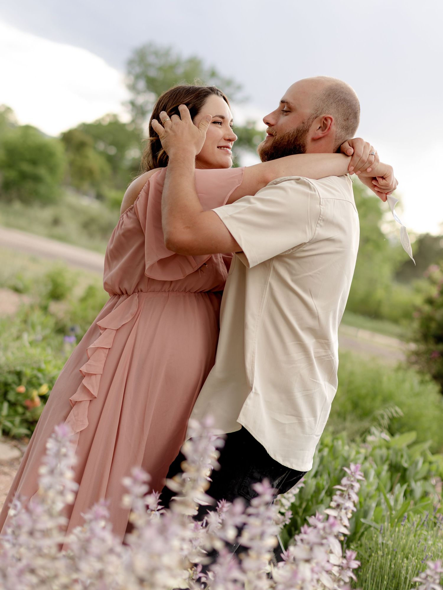 A couple embraces and smiles in a lush garden with purple wildflowers, the woman wearing a flowing pink mauve dress.