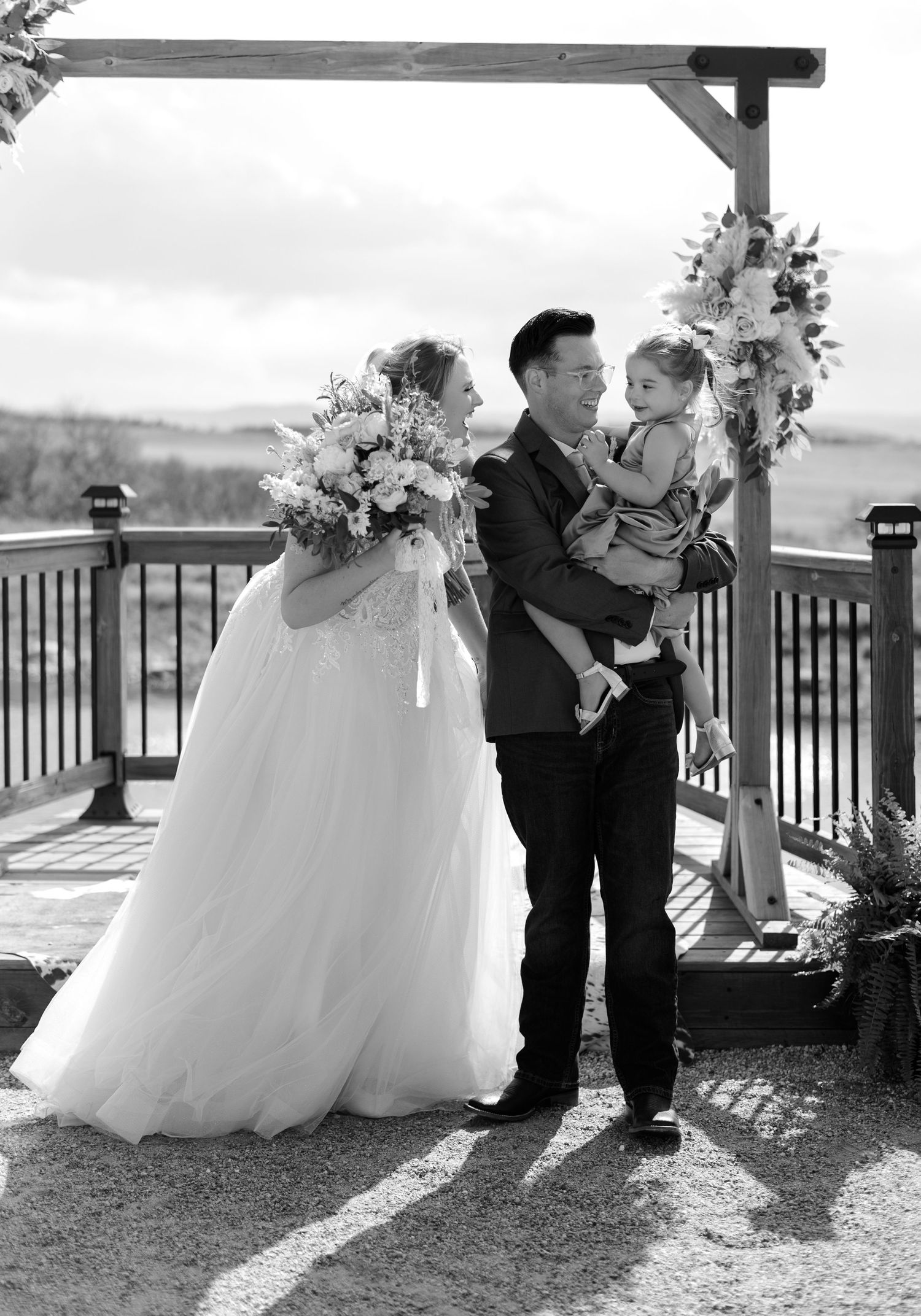 Black and white wedding photo of bride, groom, and child under floral arbor on outdoor deck.
