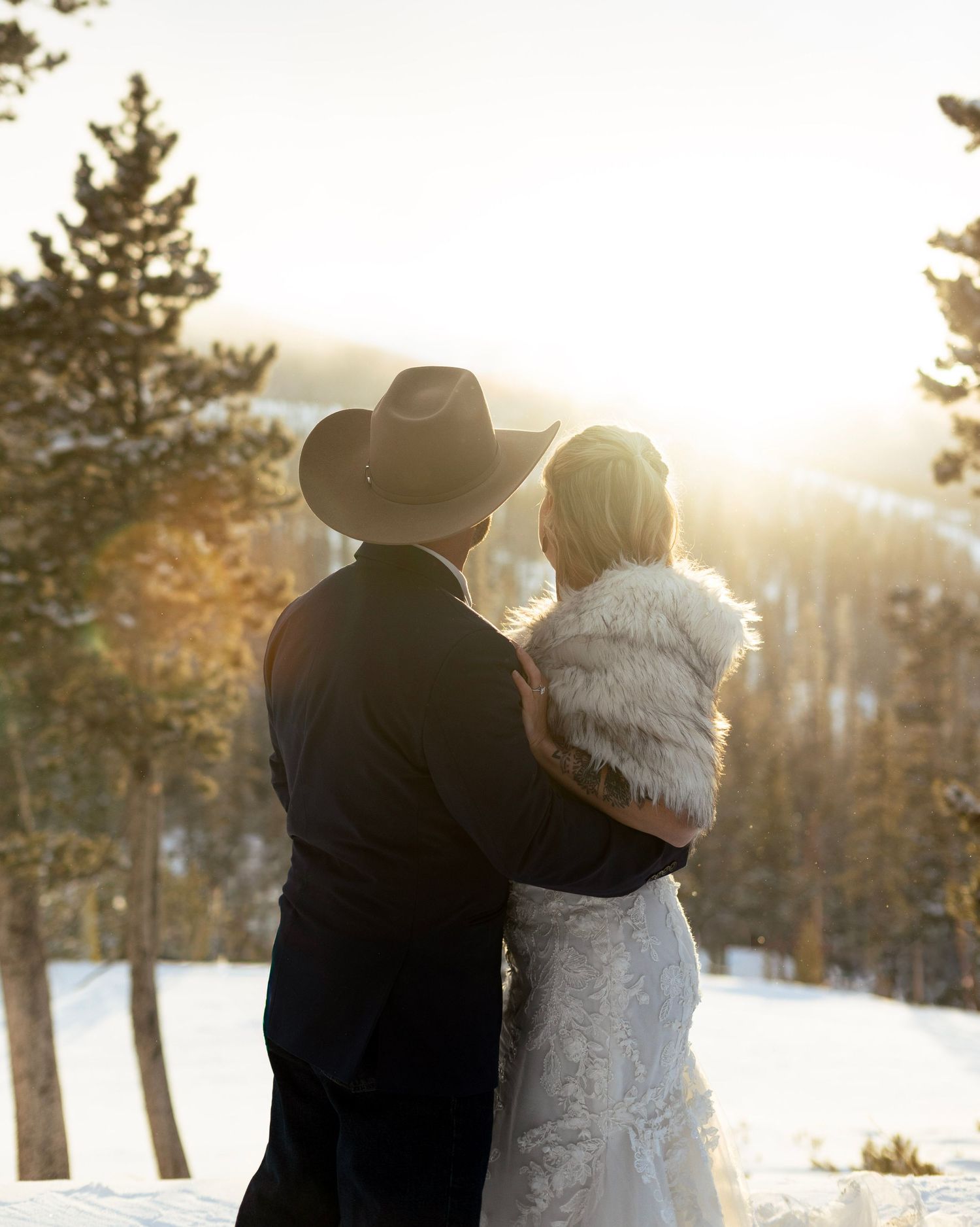 A couple in winter wedding attire embraces while watching the sunset in a snowy mountain forest setting.