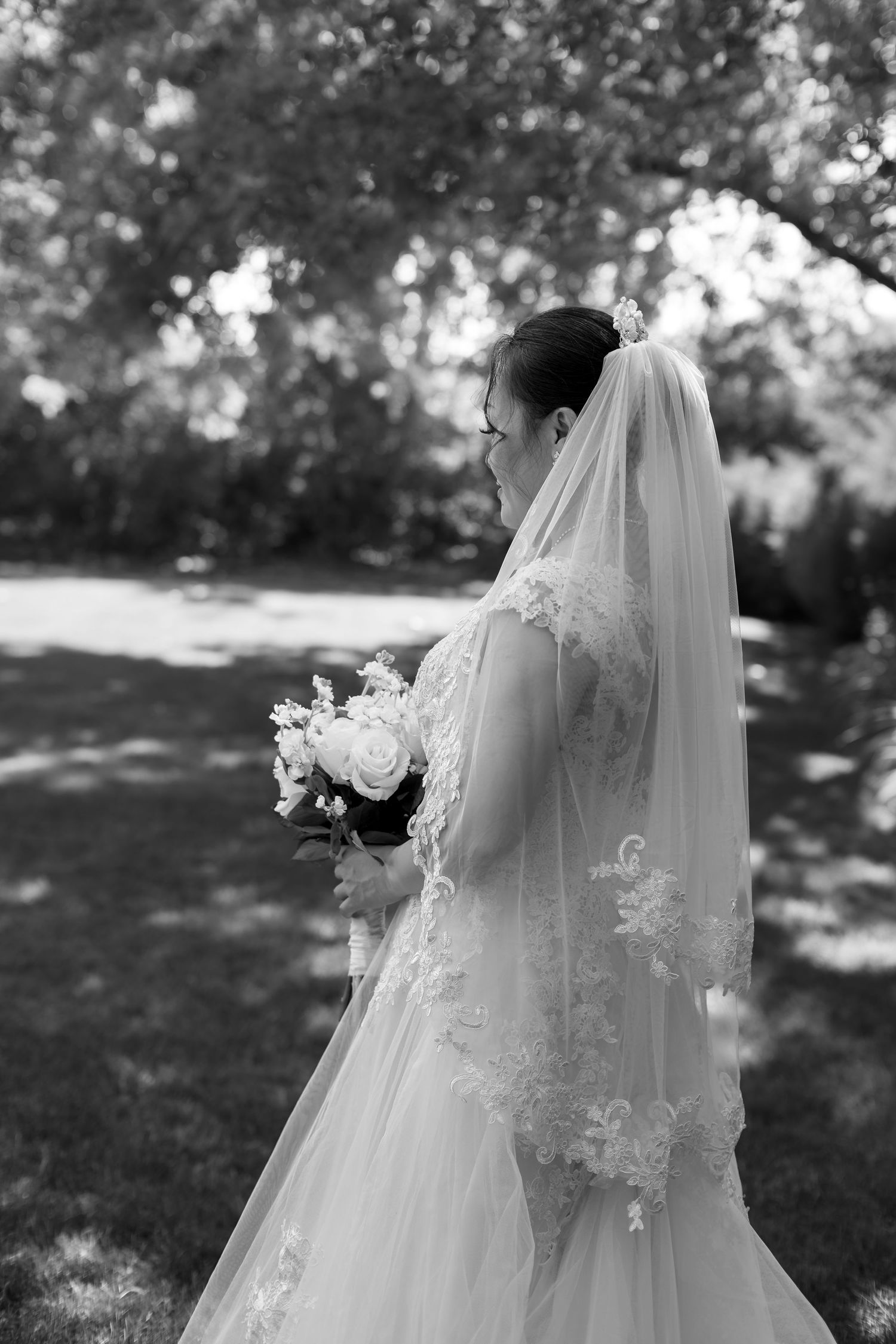 Side profile of bride in lace gown and veil holding bouquet outdoors in black and white photo.