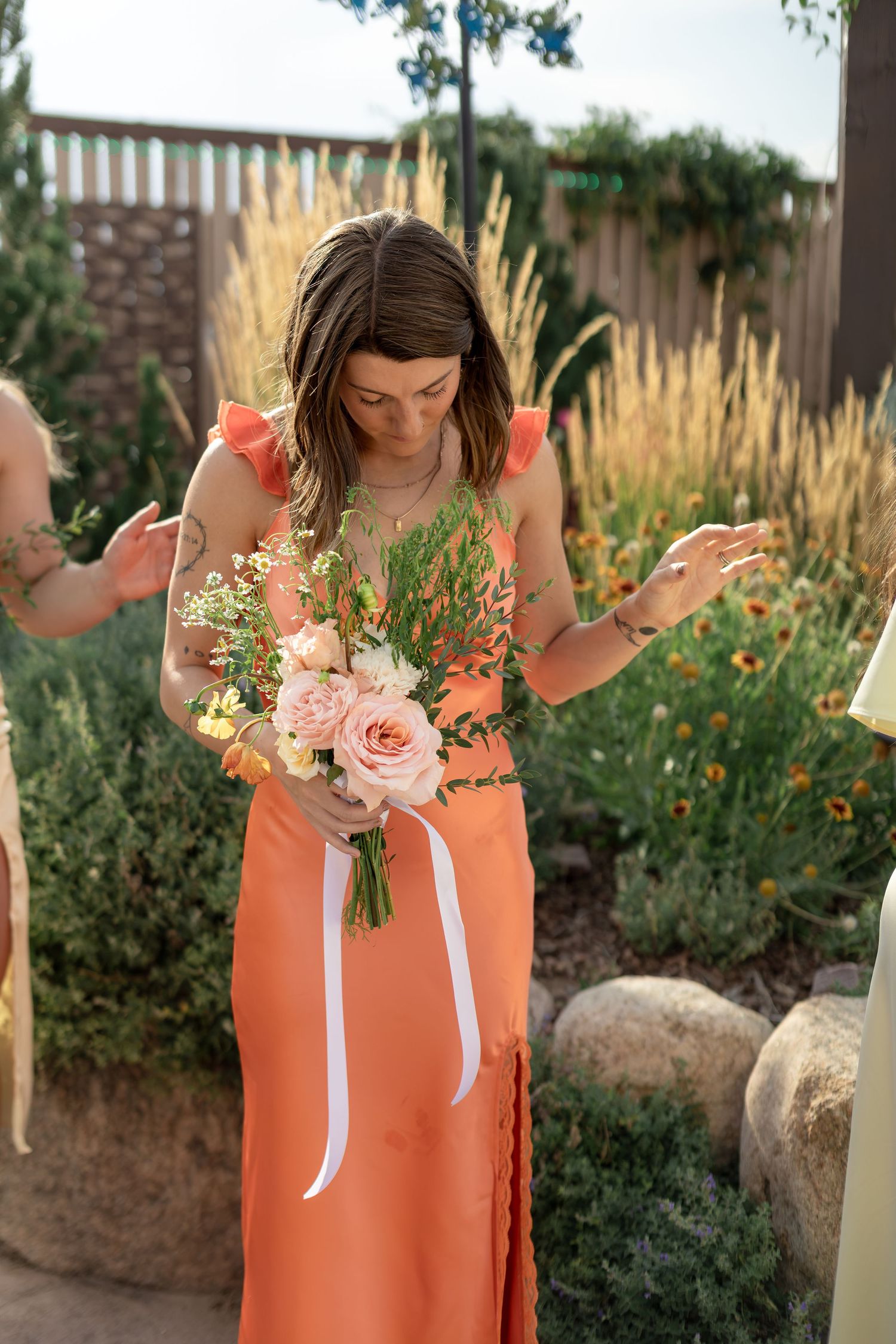 Bridesmaid in coral orange dress holding a pink rose bouquet with white ribbon at outdoor wedding.