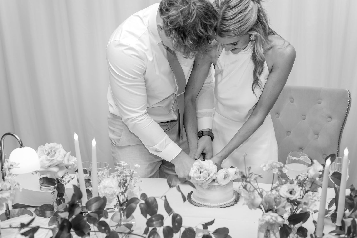 Black and white photo of bride and groom cutting wedding cake surrounded by candles and floral arrangements.