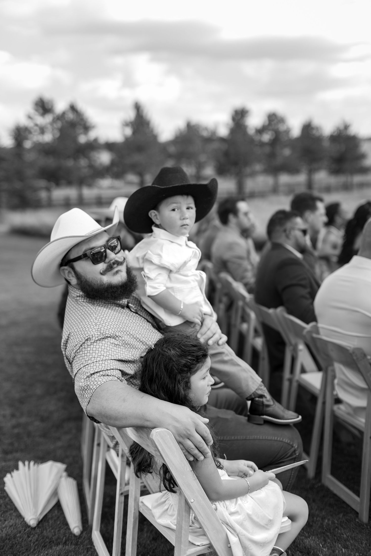 Father in cowboy hat holding young child at outdoor wedding ceremony on grassy field.