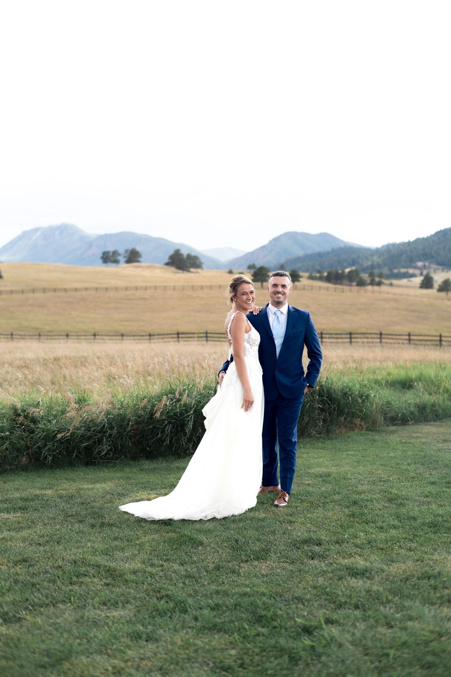 Bride and groom posing in a green meadow with golden fields and Rocky Mountain backdrop on their wedding day.
