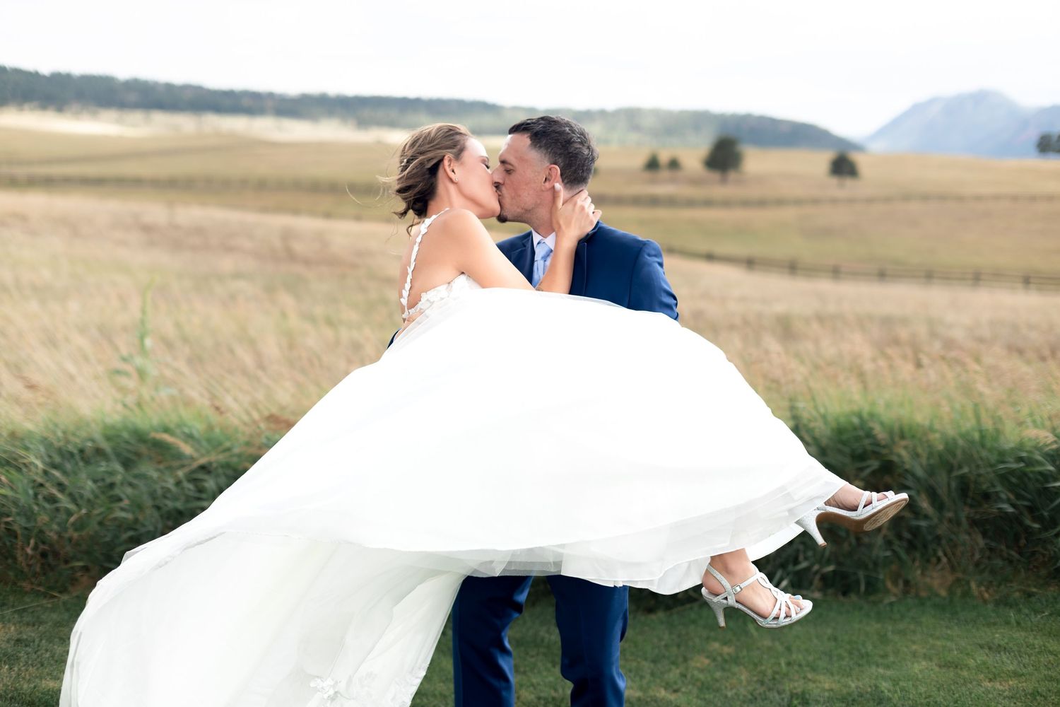 Groom lifting bride in white gown for a romantic kiss in a scenic field during outdoor wedding photo session.