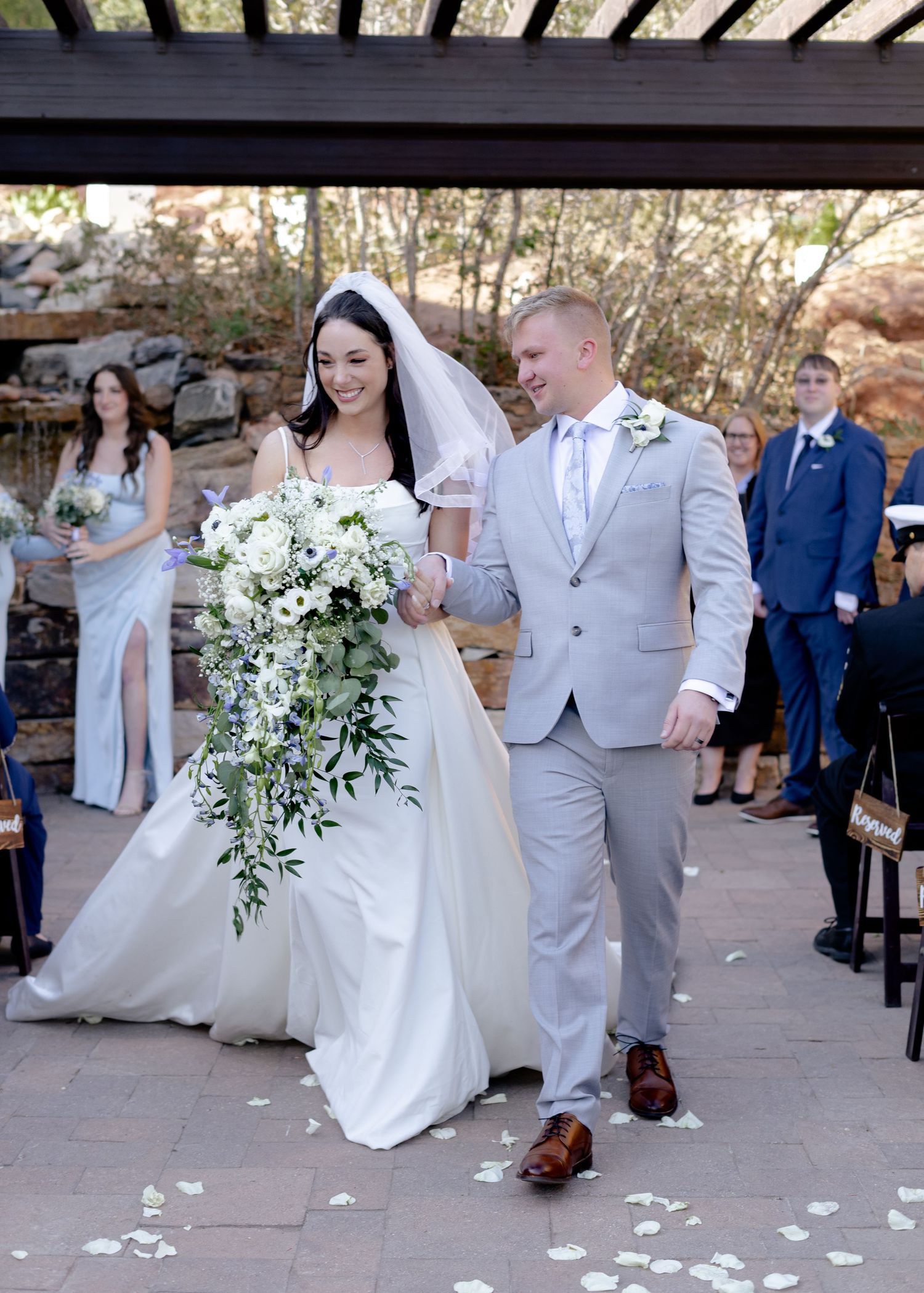 Smiling bride and groom walk down the aisle at an outdoor wedding ceremony with white floral decorations.