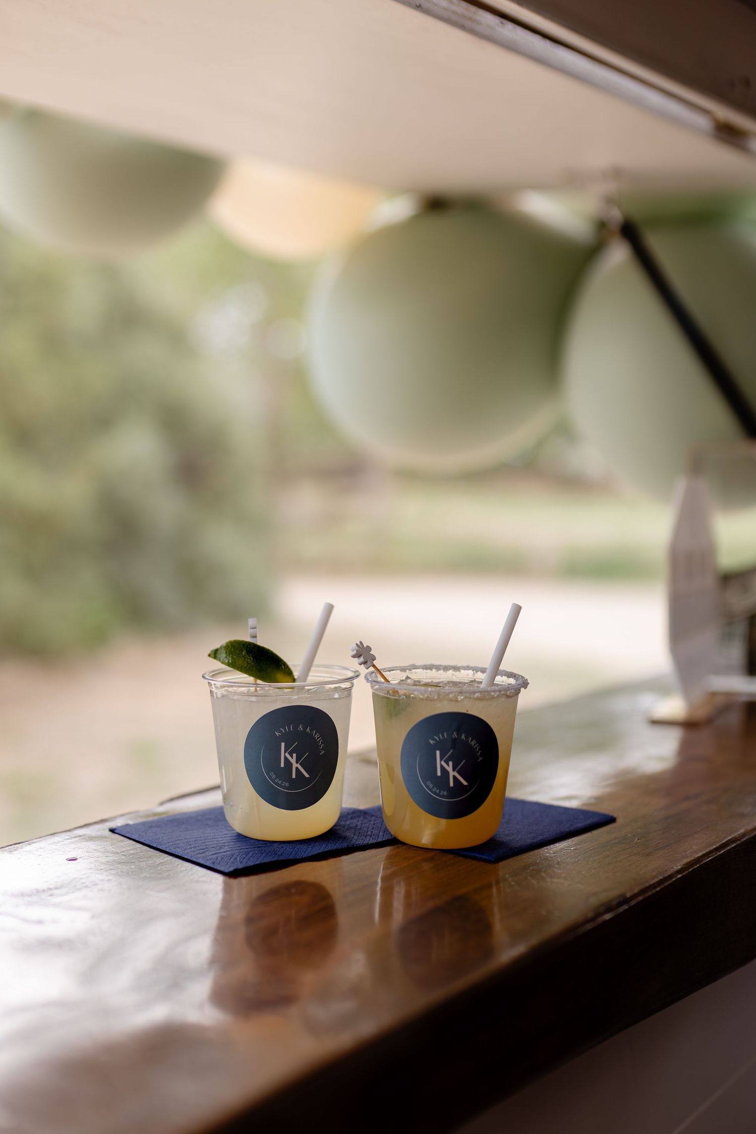 Two margarita cocktails with KK monogram logos on navy napkins at outdoor wedding bar.