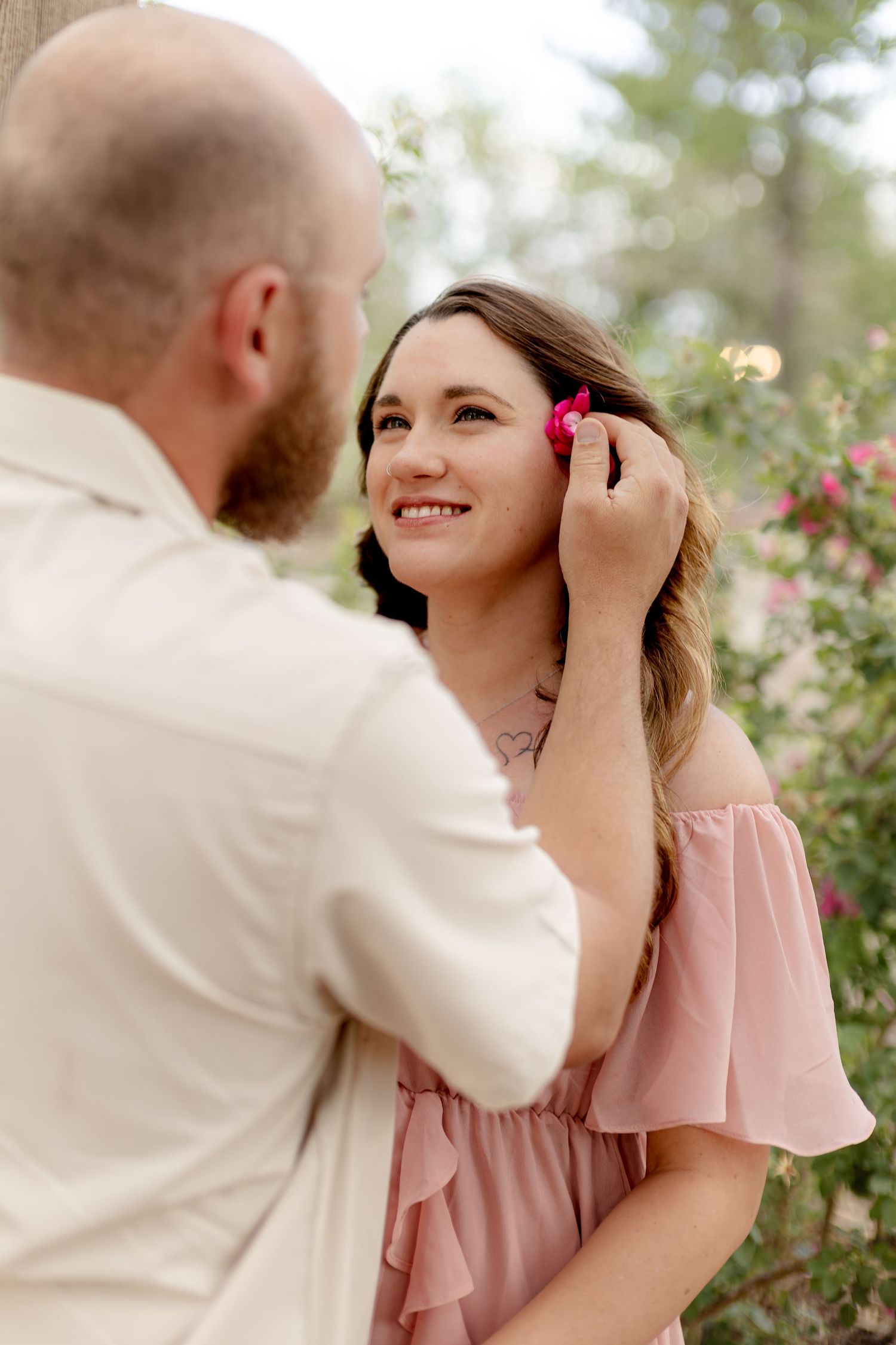 A man places a pink flower in a smiling woman's hair outdoors among blooming roses.