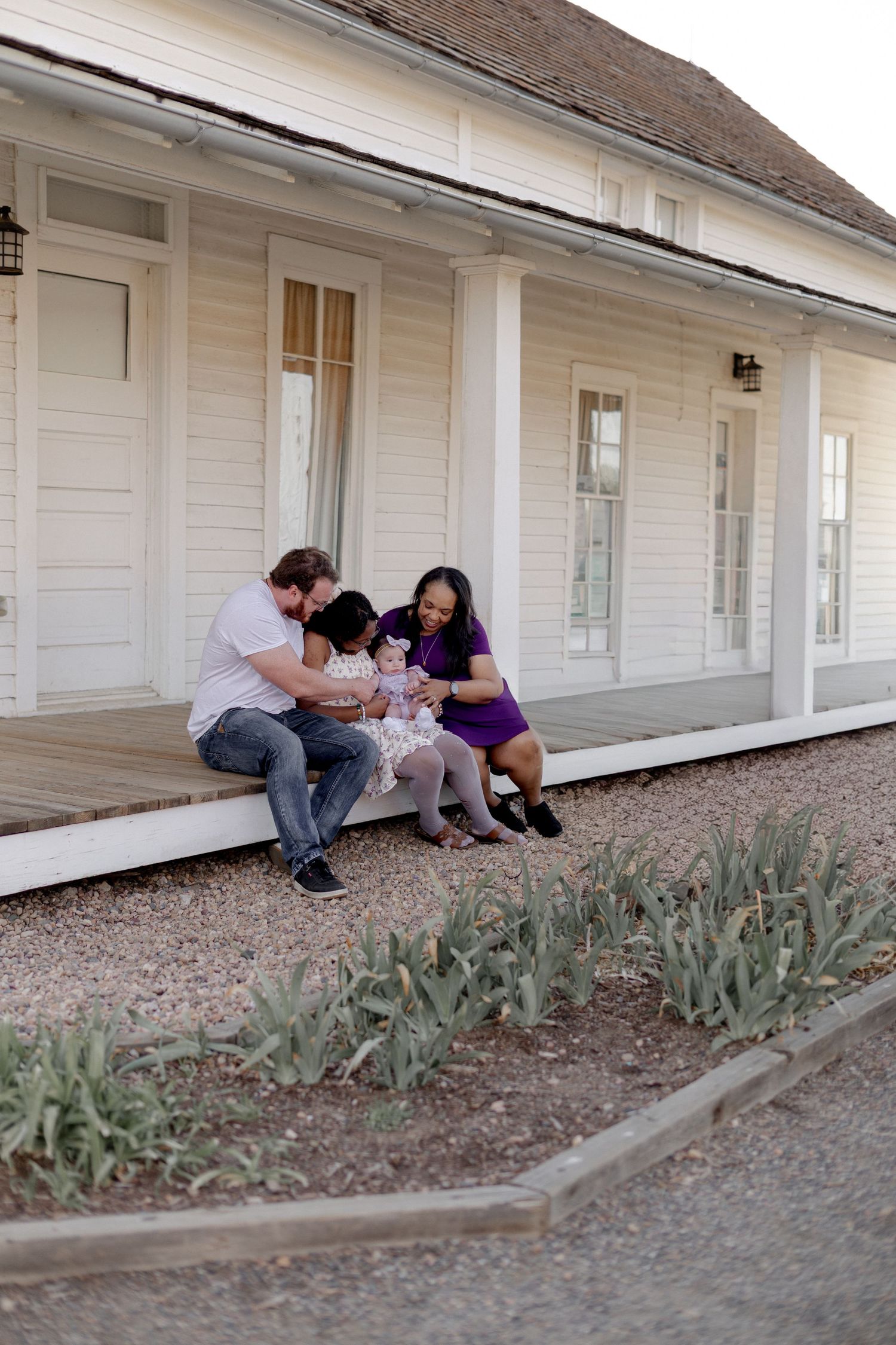 Family of three sitting on a white farmhouse porch, parents bonding with toddler in purple dress outdoors.