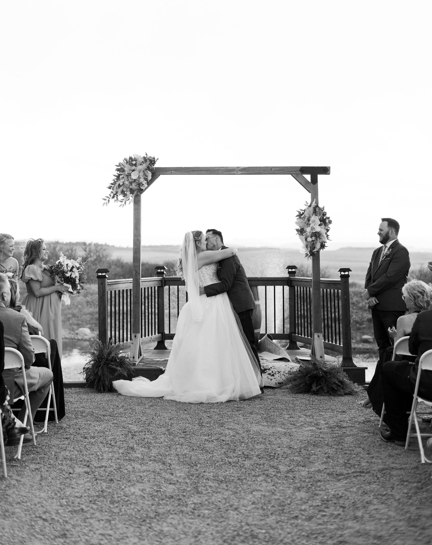 Black and white outdoor wedding ceremony with bride and groom kissing under a rustic wooden arch.
