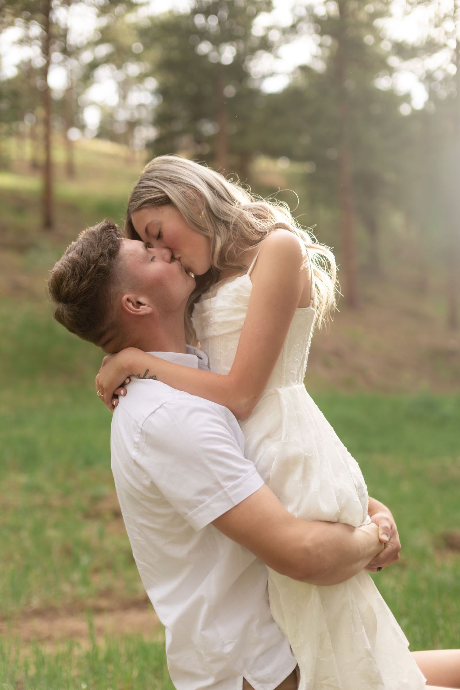 Couple sharing a romantic kiss outdoors, man lifting woman in white dress, surrounded by lush green trees.