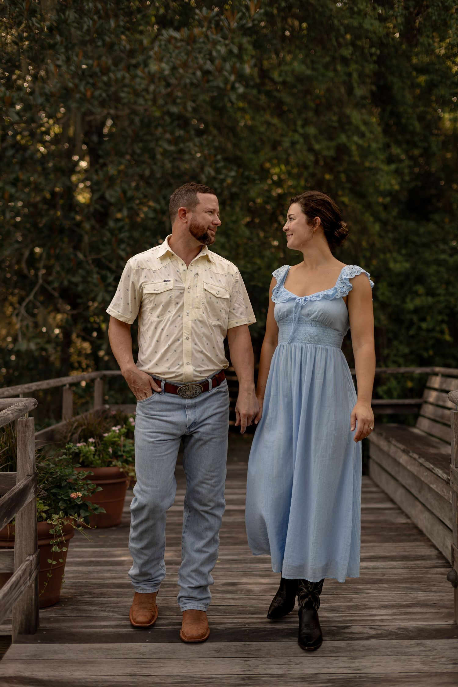 Couple holding hands and walking on a wooden bridge surrounded by greenery in a garden setting.