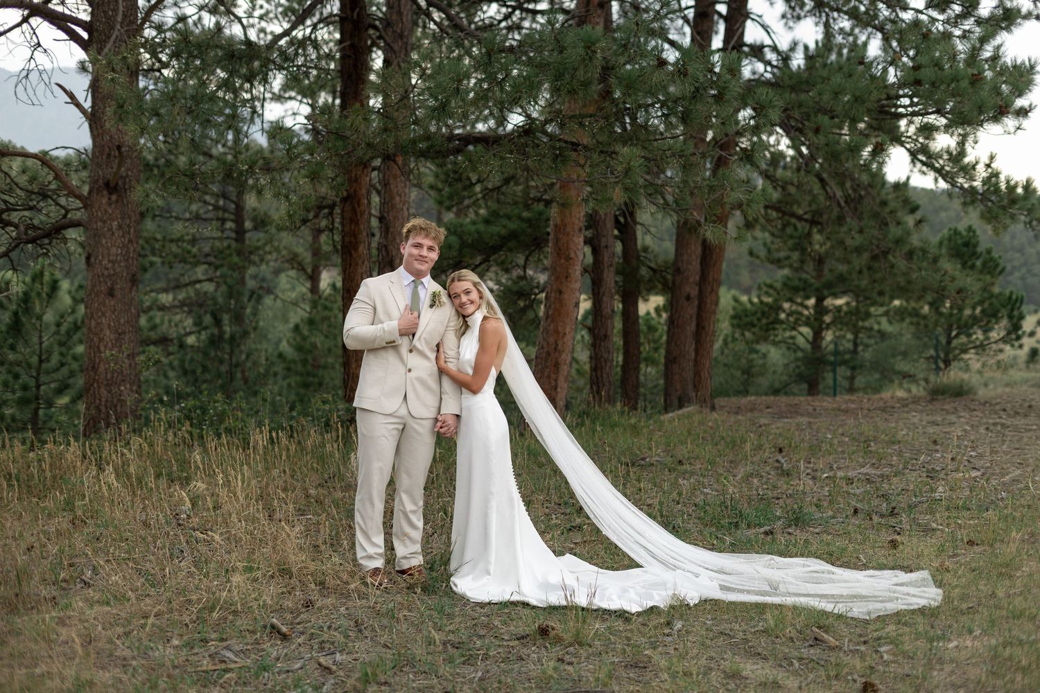 Bride and groom posing together outdoors among tall pine trees, bride in elegant white gown with cathedral veil.