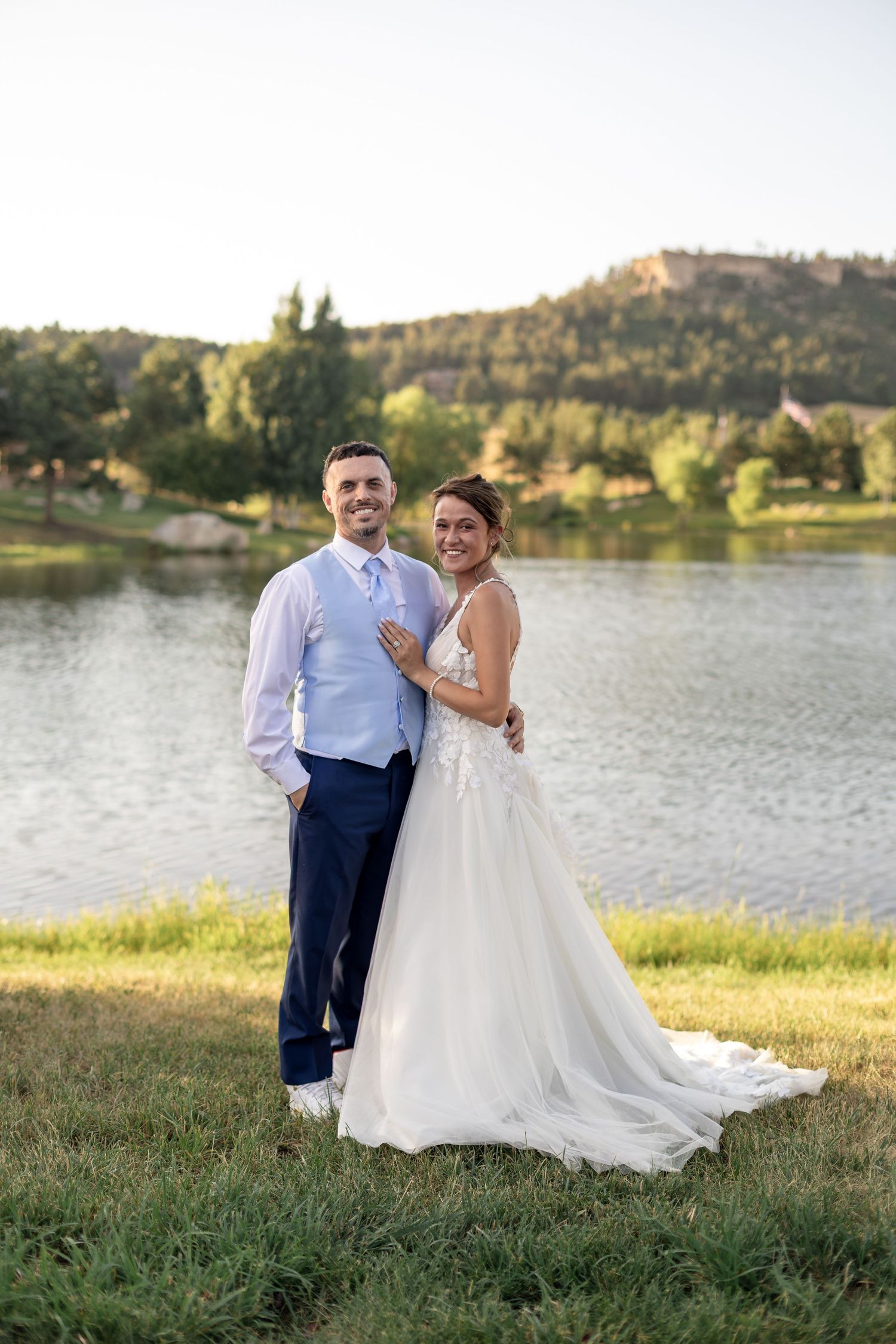 Bride and groom pose together smiling by a lake with mountain backdrop at golden hour.
