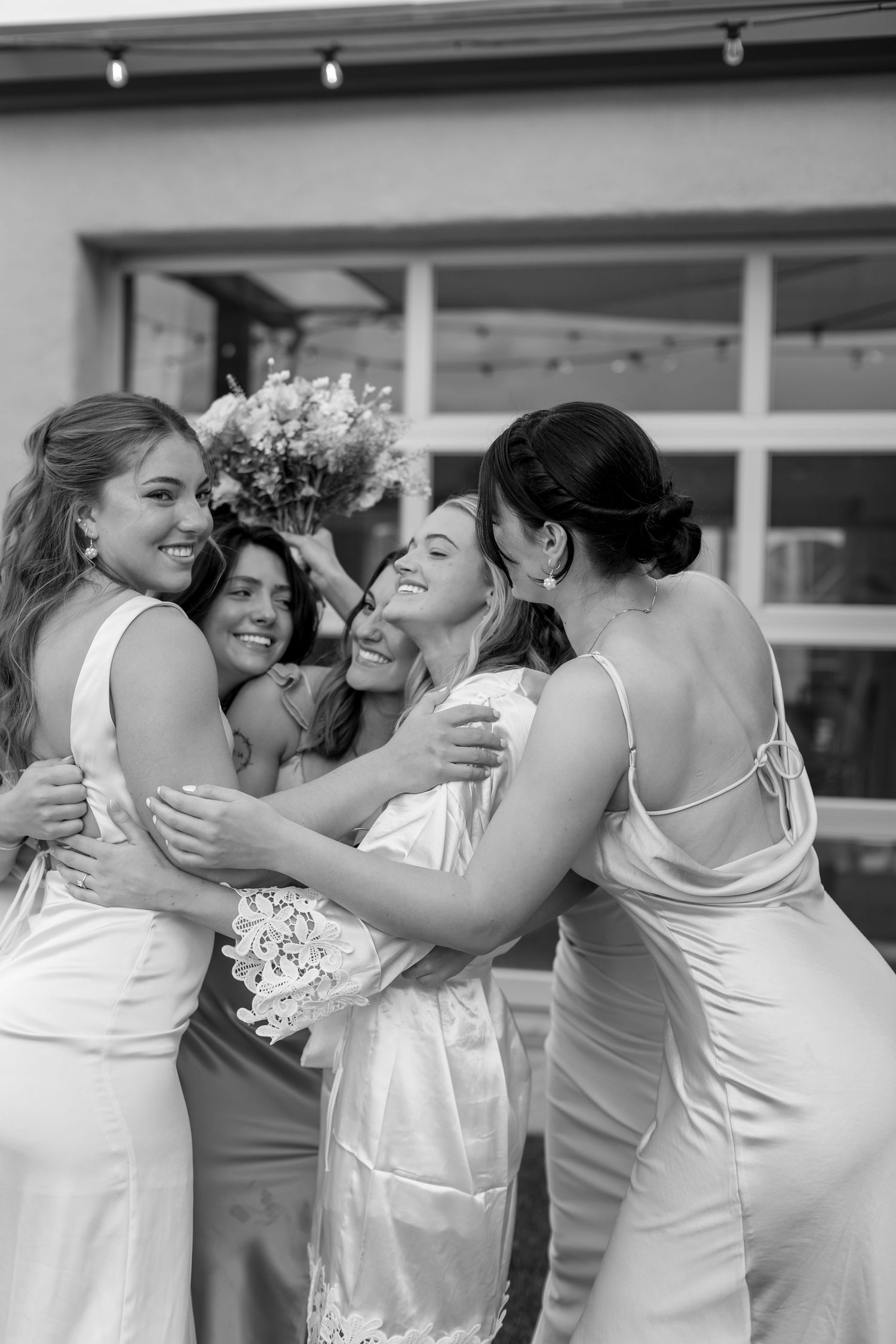 Black and white photo of a bride in a white gown hugging her bridesmaids while holding a bouquet outdoors.