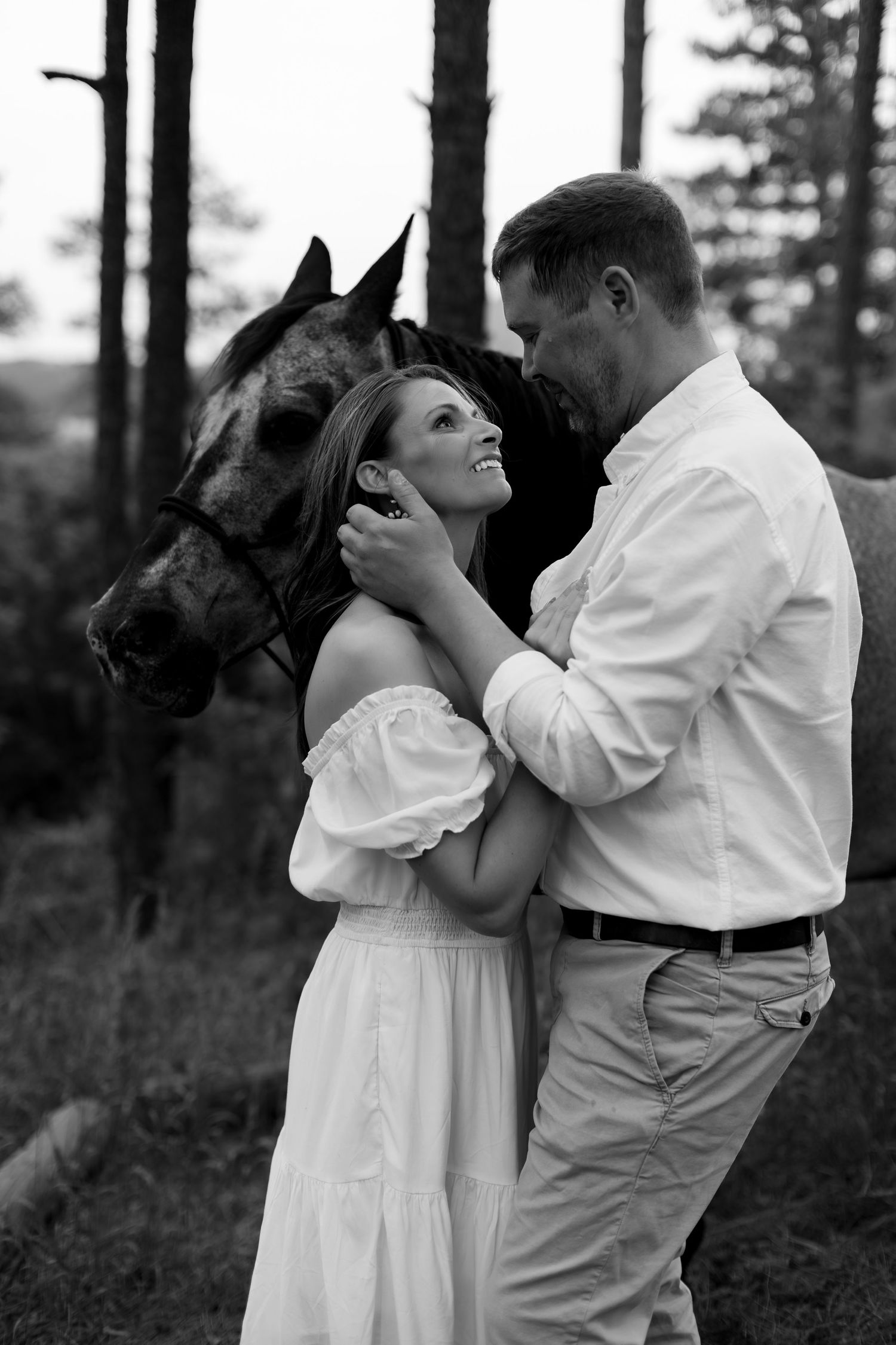 Black and white photo of a romantic couple embracing in a forest with a horse standing behind them.