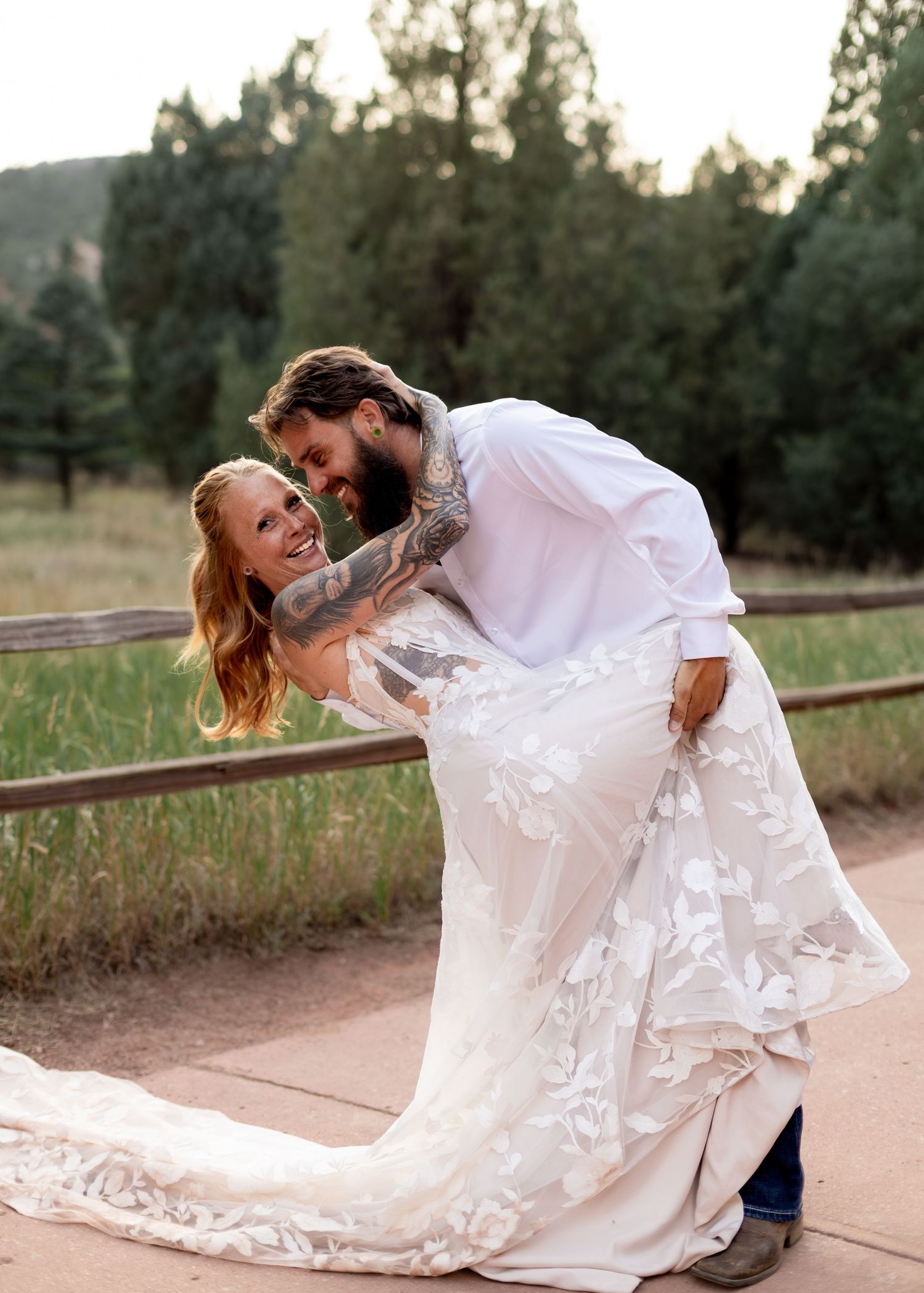 Groom dipping laughing bride in floral lace wedding gown outdoors at rustic Colorado wedding venue.