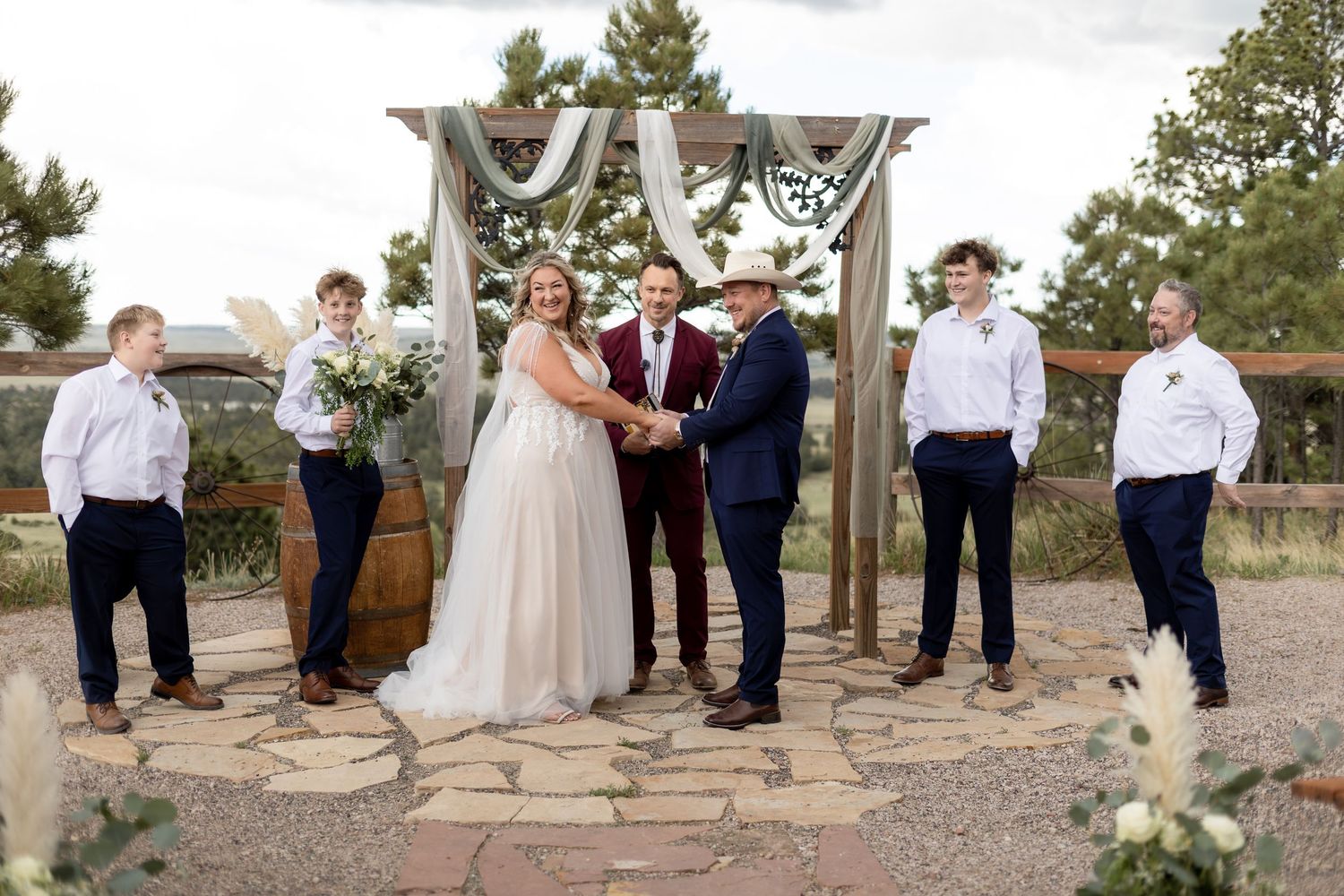 Outdoor wedding ceremony with bride and groom holding hands at rustic wooden arch, flanked by groomsmen in navy suits.