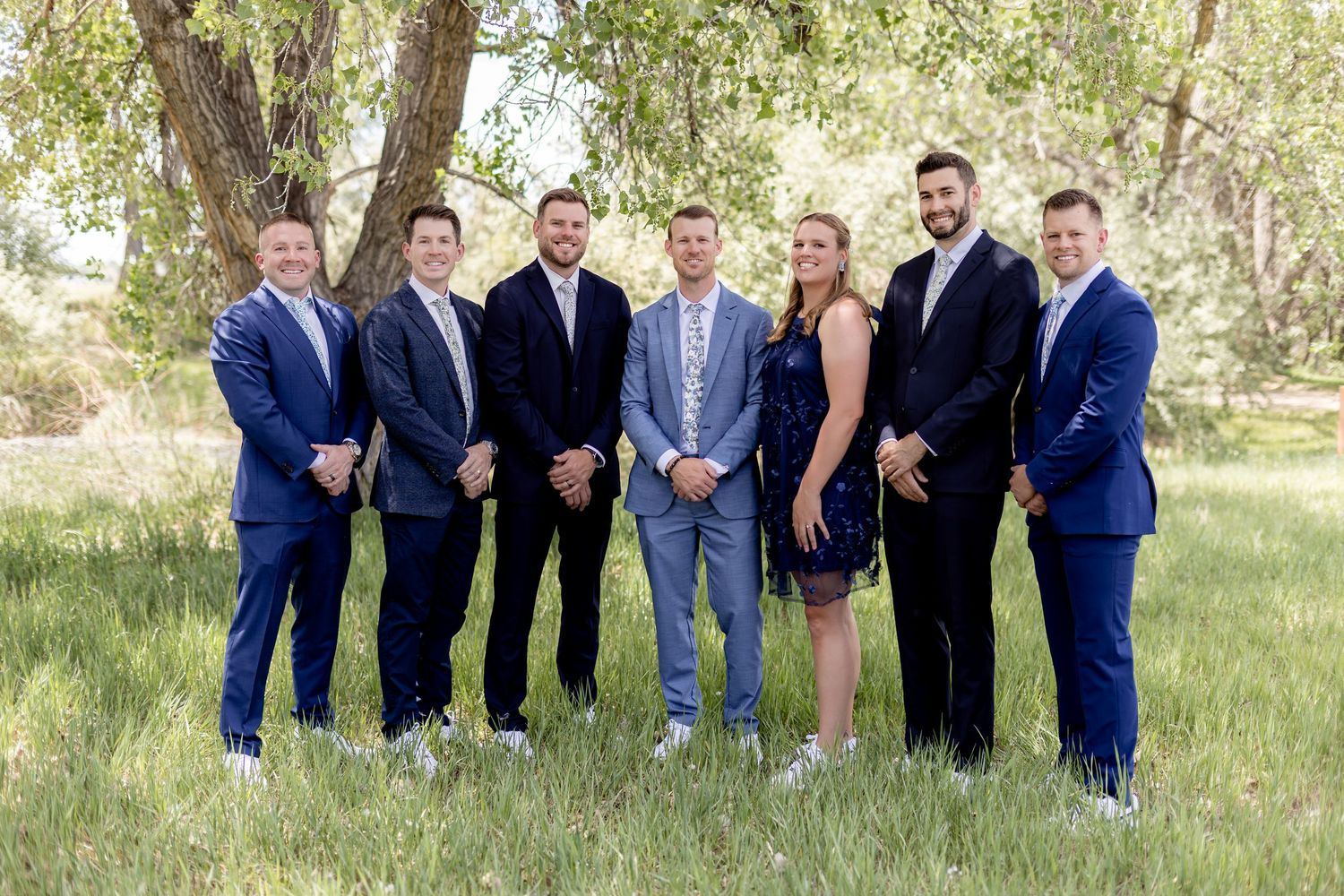 Group of seven professionals in blue suits posing outdoors in a lush green field near a large tree.