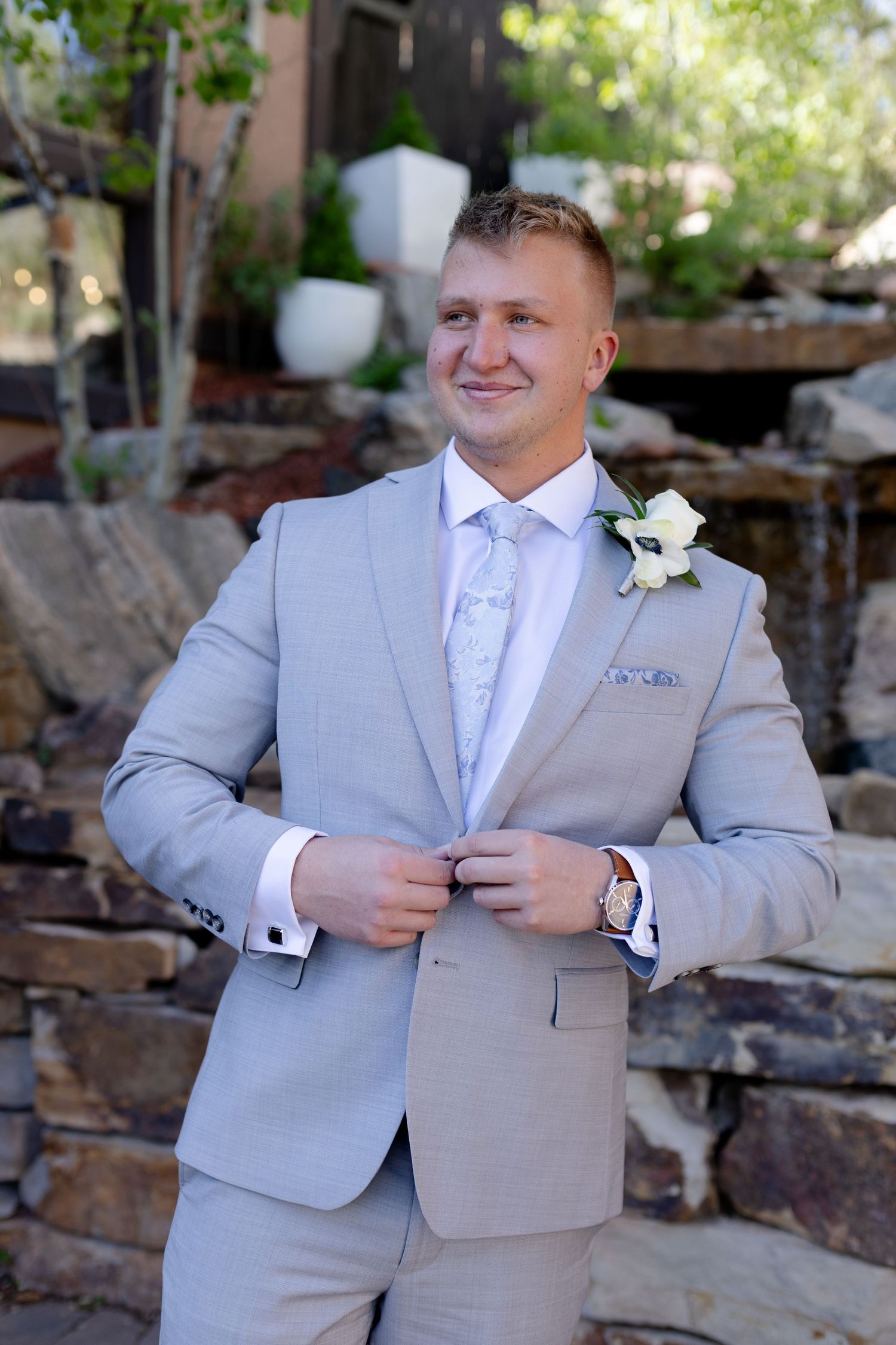 Dapper groom in light gray suit with white boutonniere adjusting jacket near stone waterfall.