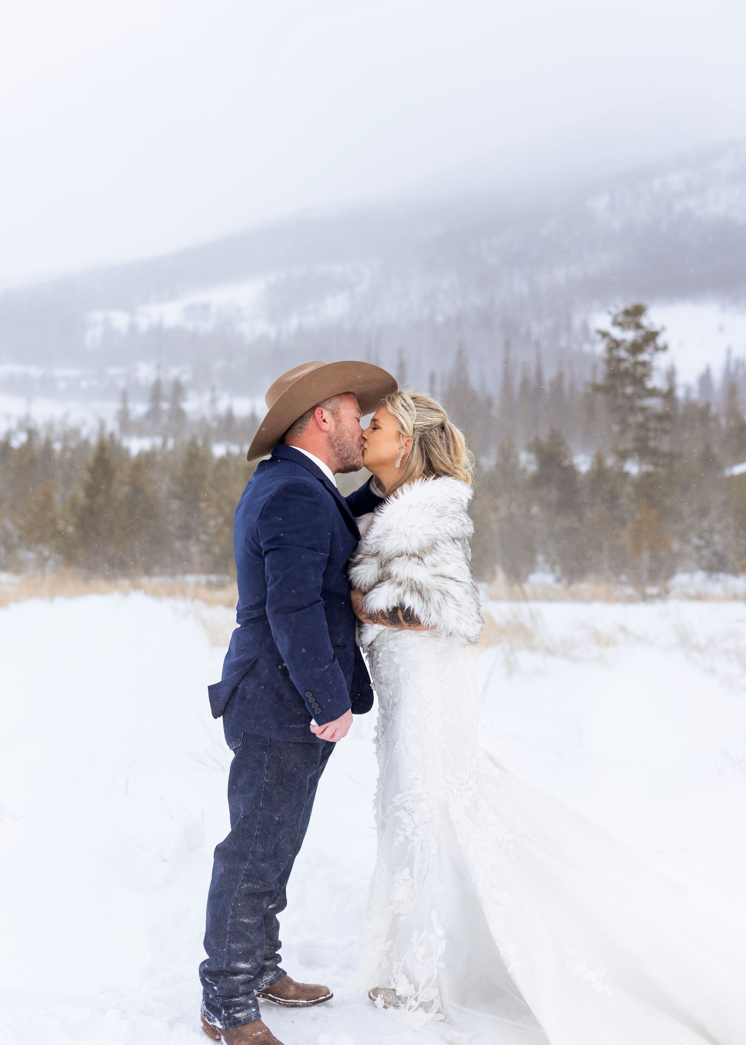 A romantic winter wedding scene with a couple embracing in the snow with mountains and evergreen trees in the background.