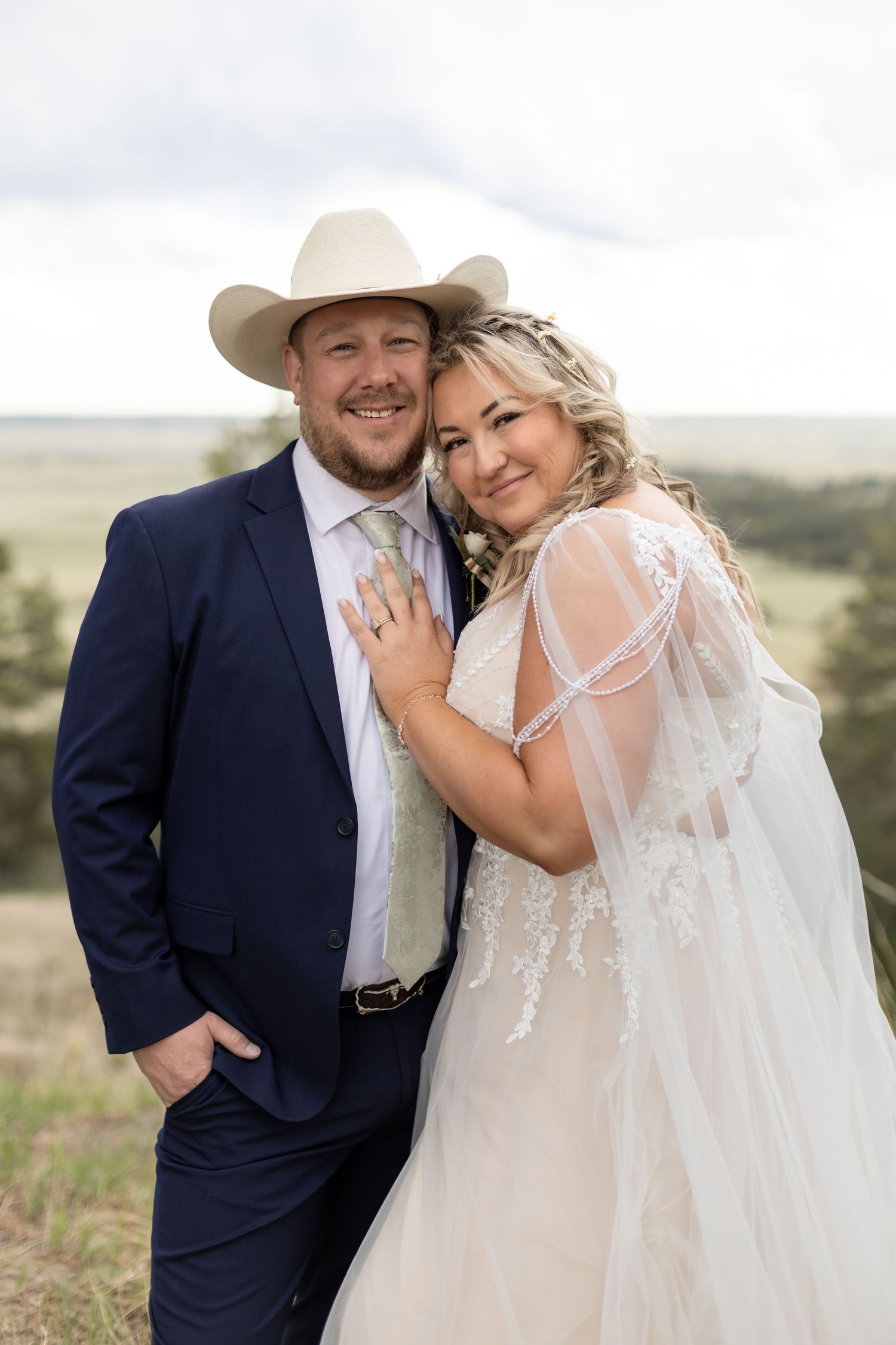 A smiling groom in a navy suit and cowboy hat poses with his bride in a lace wedding dress outdoors.