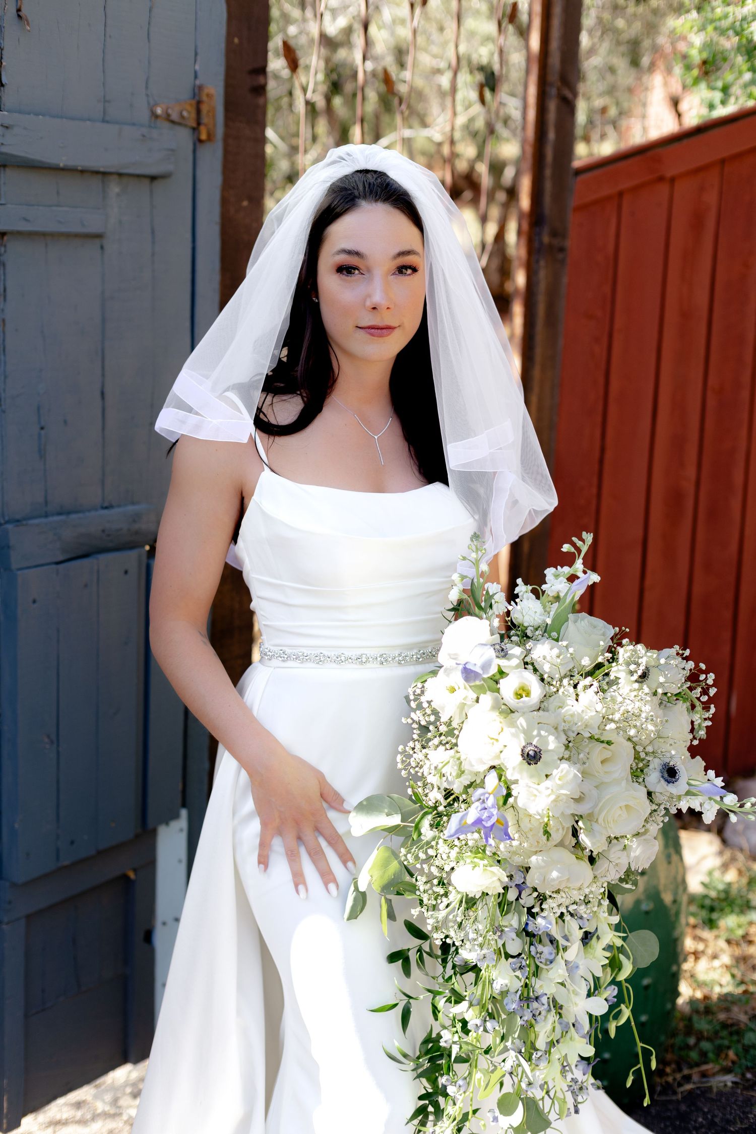Bride in white gown and veil holding cascading white floral bouquet outdoors.