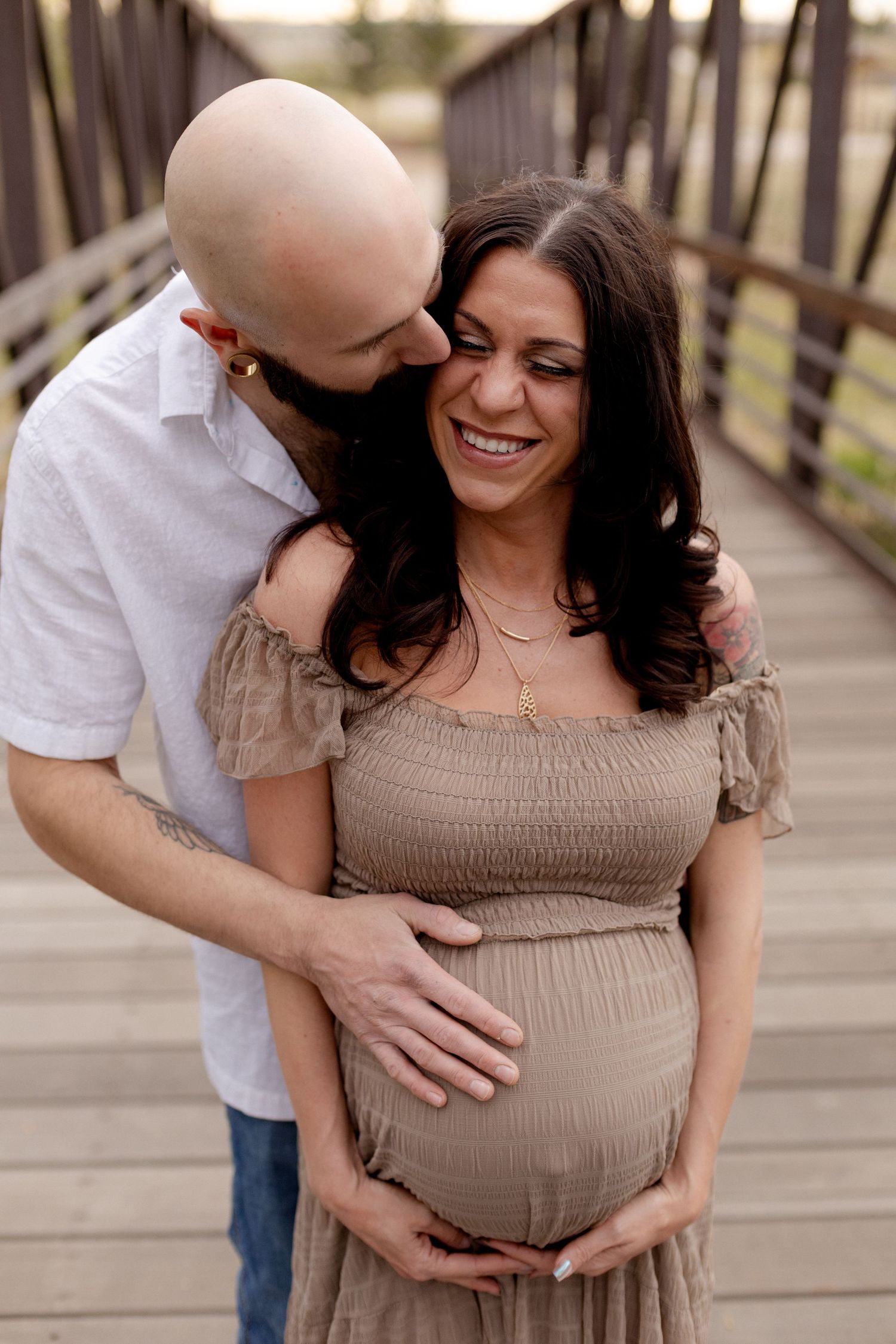 Smiling pregnant woman in tan dress embraced by bald bearded man on wooden bridge during maternity photo session.