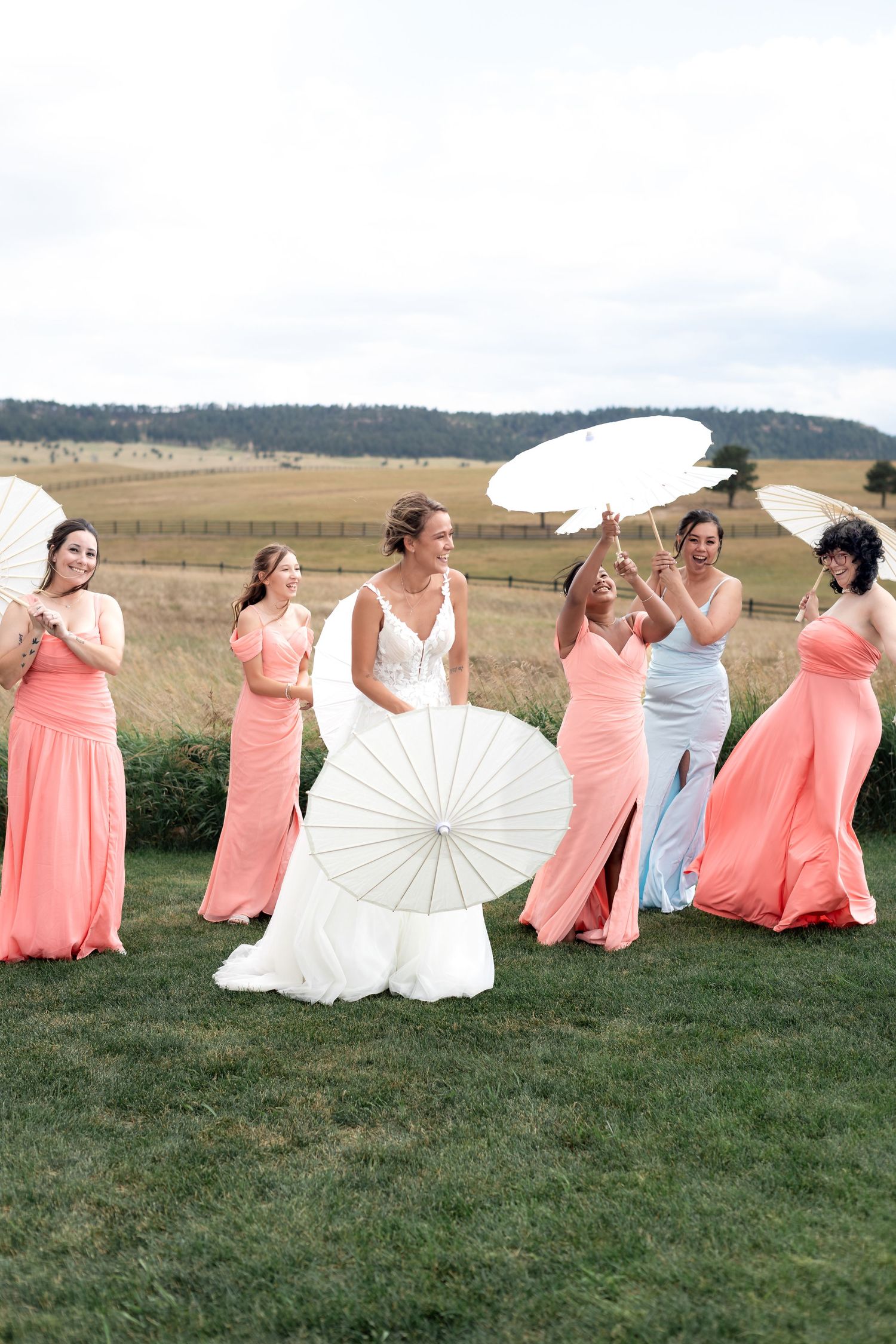Bride in white gown holds parasol with bridesmaids in coral dresses holding white parasols on green lawn.