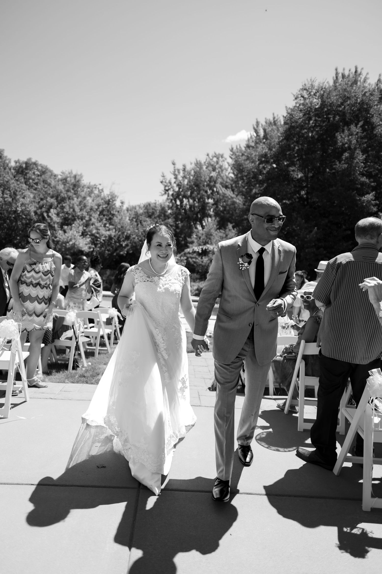Black and white photo of bride and groom walking down outdoor wedding aisle holding hands on sunny day.