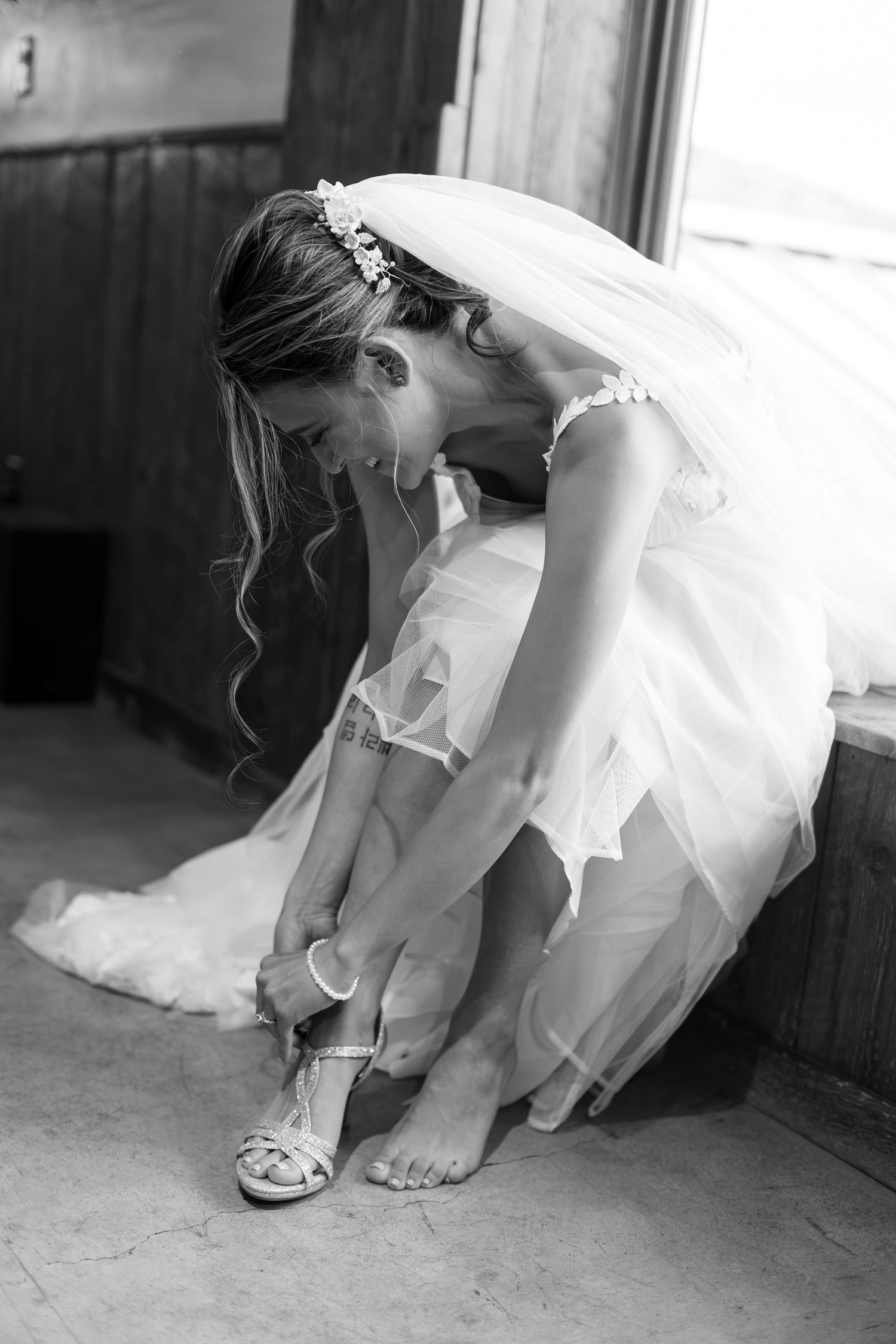 Black and white photo of a bride in a veil adjusting her wedding heels while seated.
